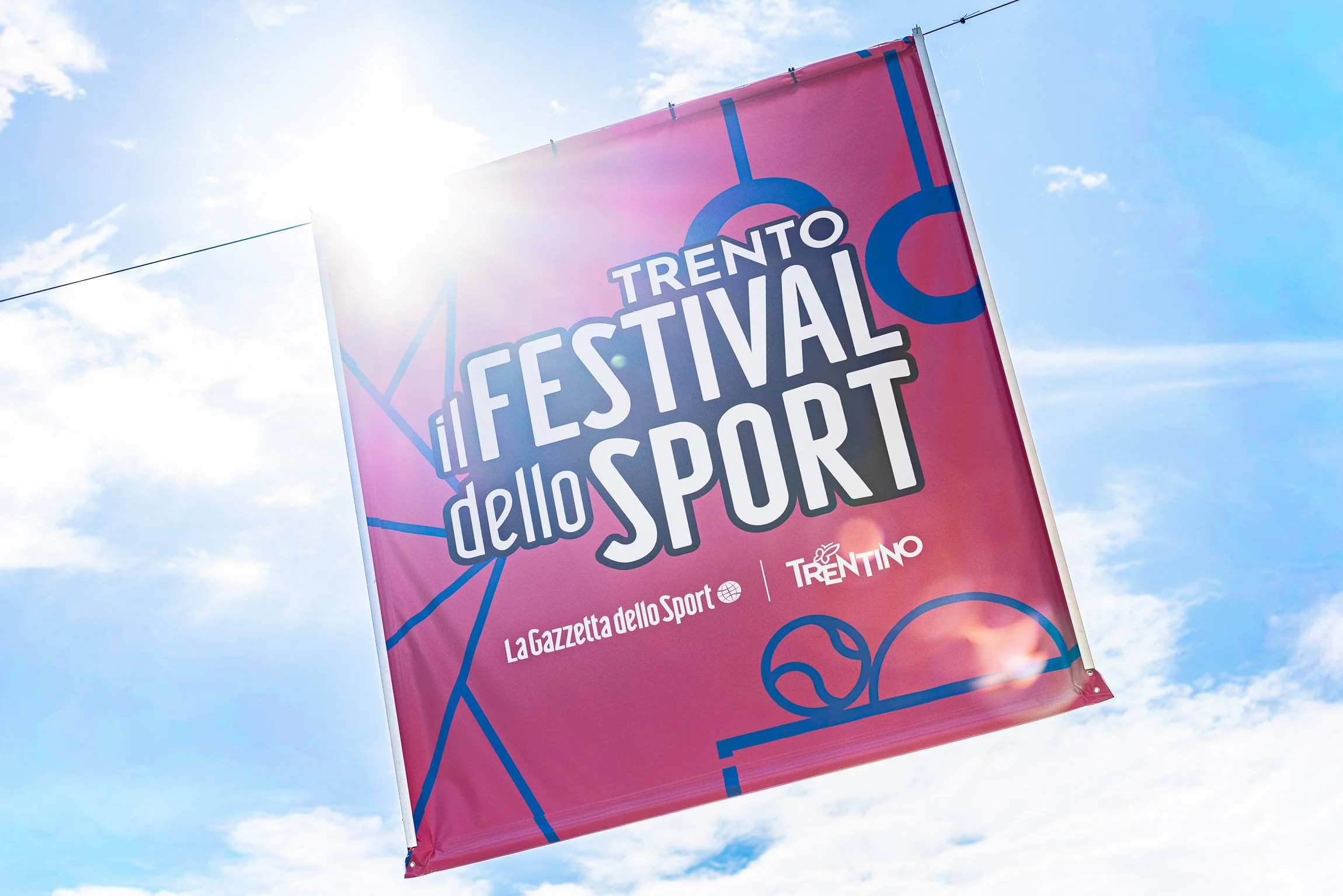 Bandiera pubblicitaria del festival dello sport a Trento con sfondo di cielo e nuvole.

© Alessandro Holneider