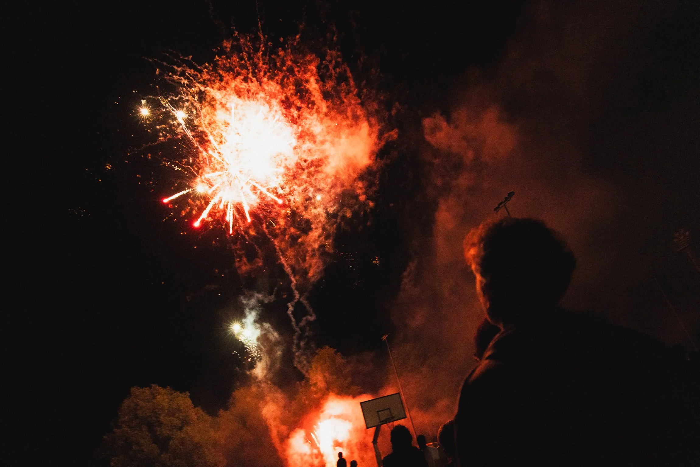 Festa con fuochi d'artificio al notte, persone che guardano il cielo e un campo da basket sullo sfondo.

© Alessandro Holneider