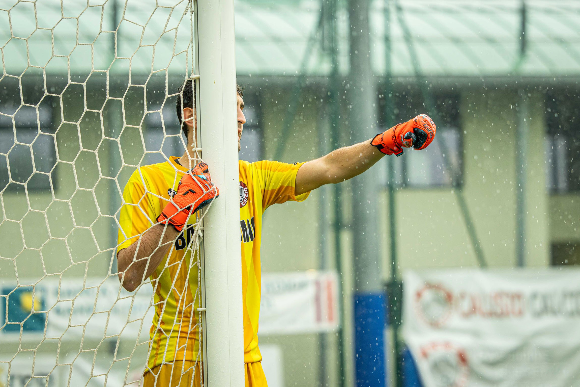 ASD Calisio Calcio. Portiere di calcio rosa in piedi dietro la rete con impermeabile arancione, che si sta bagnando sotto la pioggia, durante una partita.

© Alessandro Holneider
