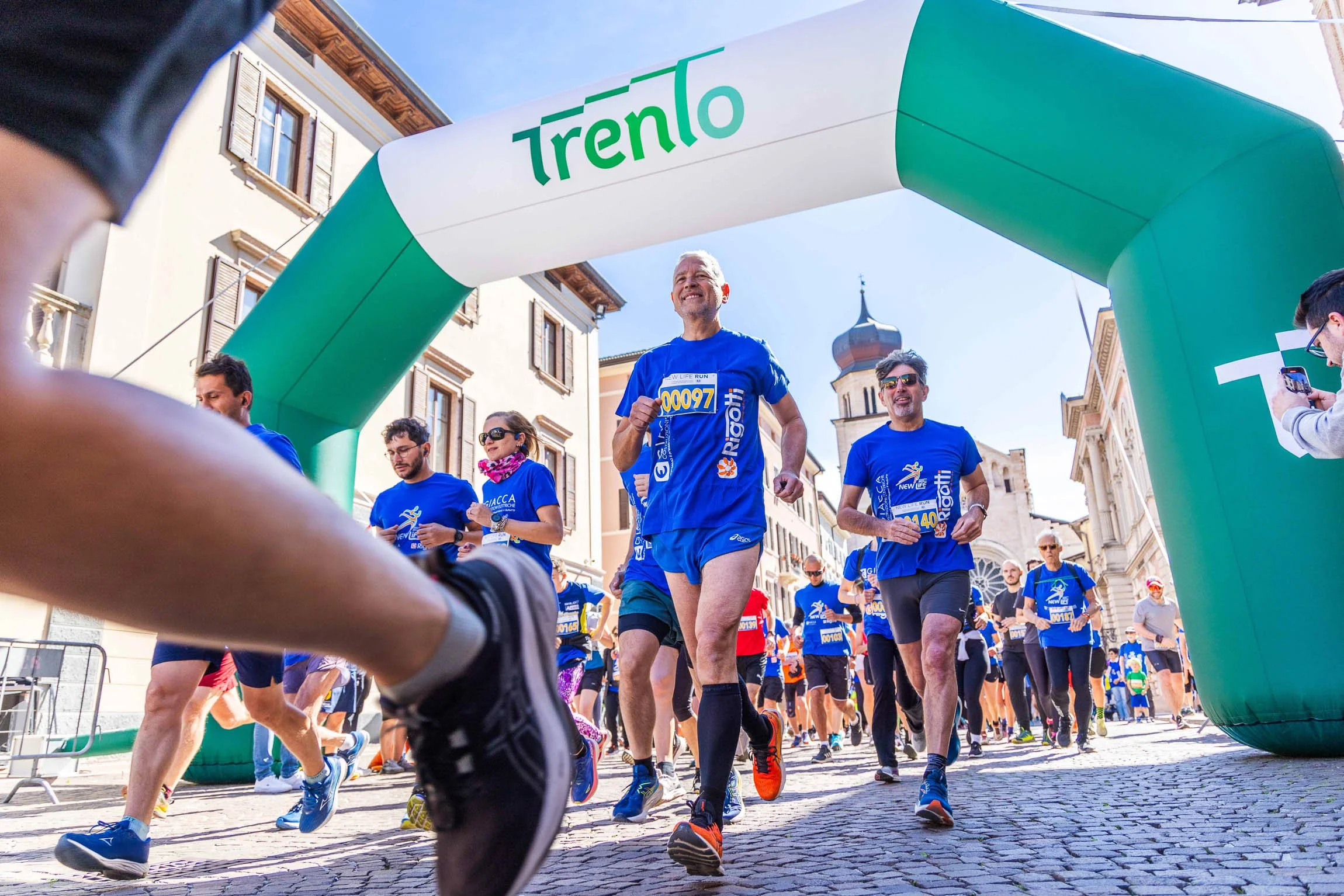 Corsa benefica in via verdi, trento. Maratona in corso con corridori in abbigliamento sportivo che attraversano un arco gonfiabile verde con la scritta "Trento" in un centro storico con edifici storici e una chiesa con torre.

© Alessandro Holneider