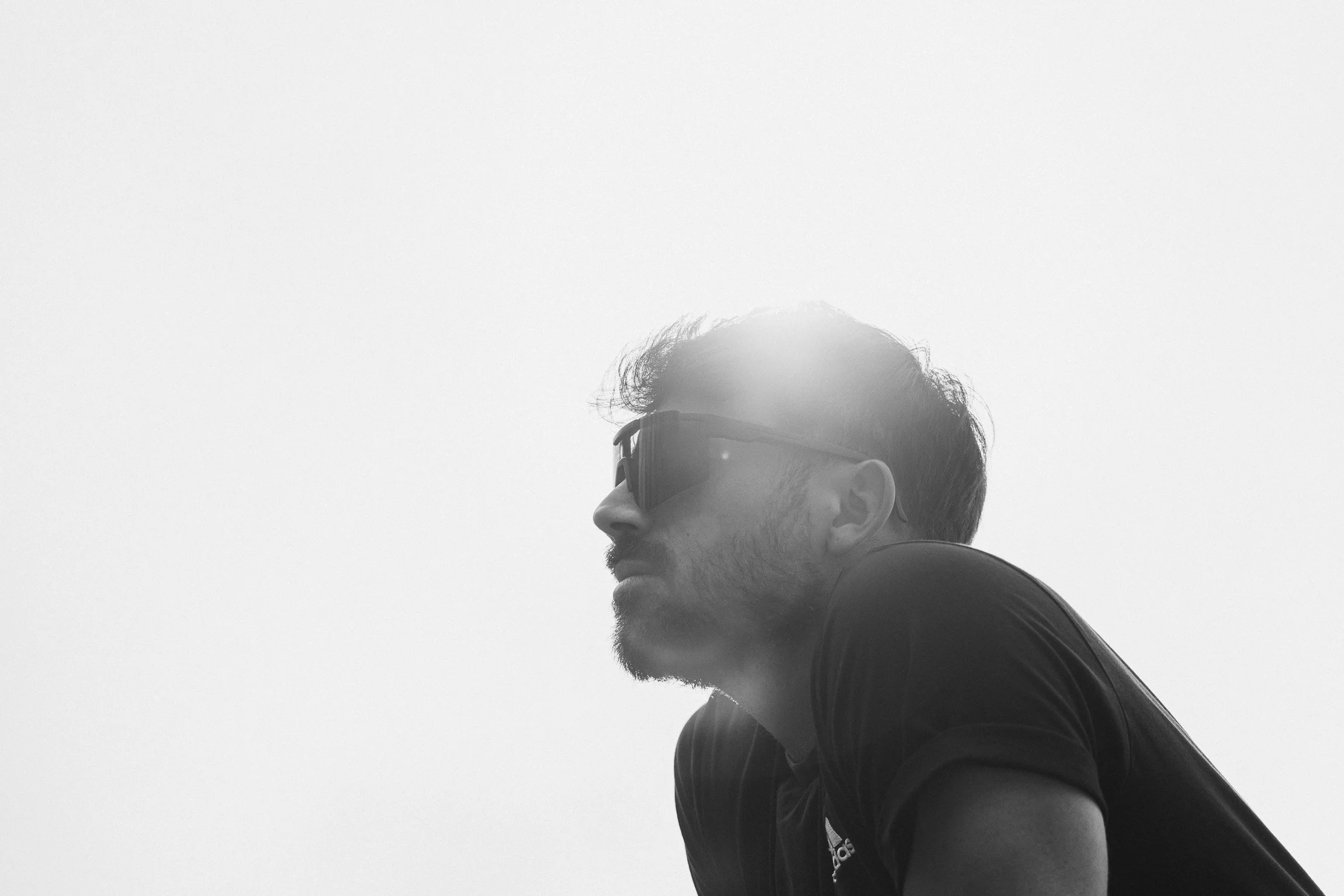 Uomo con barba e capelli corti, indossa occhiali da sole e una maglietta nera, si trova all'aperto con sfondo bianco e luce solare che crea un alone di luce sopra la testa.

© Alessandro Holneider