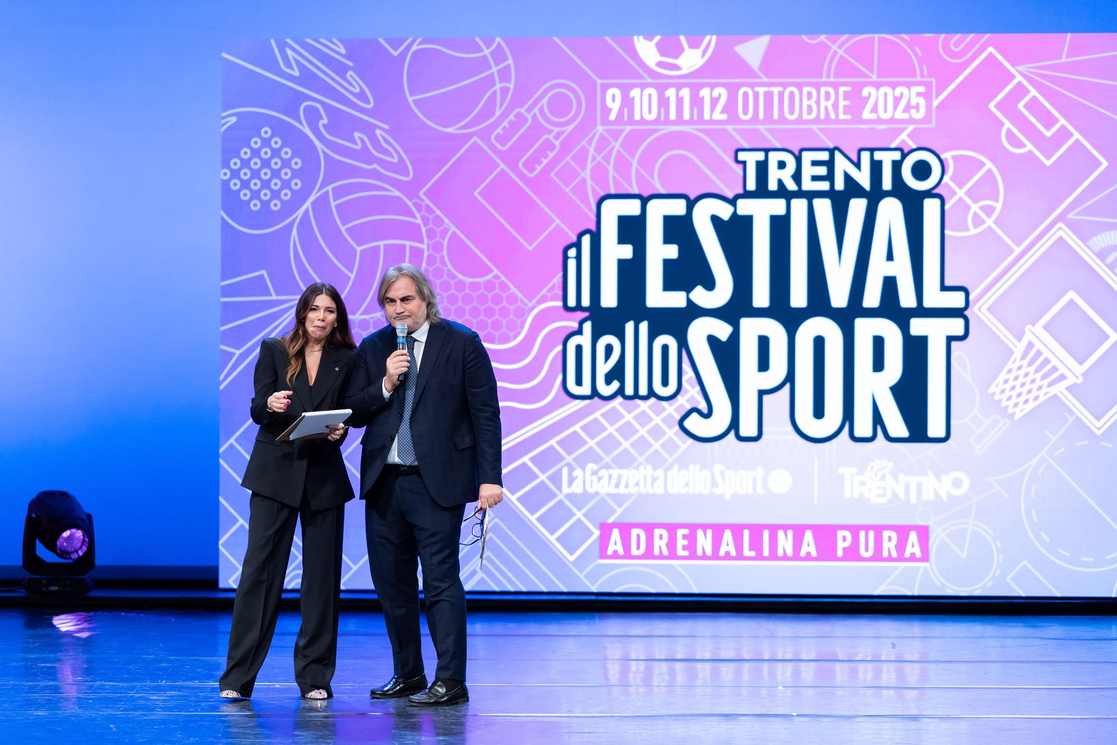Federica Masolin e Pierluigi Pardo al Festival dello Sport di Trento. Due persone sul palco di fronte a uno schermo con il logo del Festival dello Sport 2025 di Trento, uno con un microfono in mano e l'altra con un tablet. Sullo sfondo, scritte e dis
