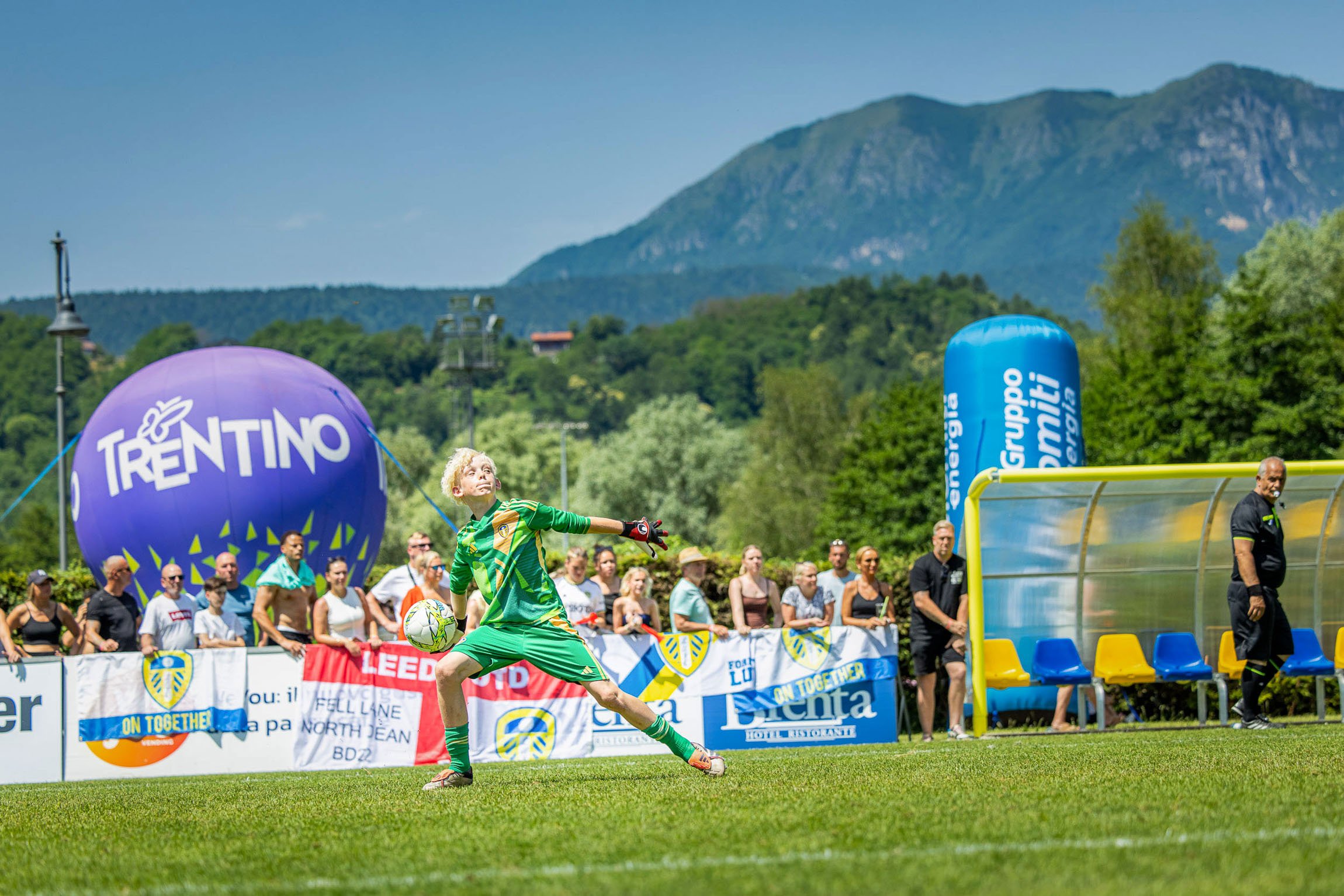 Torneo Internazionale Pulcino d'Oro, Levico Terme - Trentino. Un giocatore di calcio, indossa una divisa verde, sta calciando un pallone durante una partita sulla coppa del Trentino, con pubblico e montagna sullo sfondo.

© Alessandro Holneider