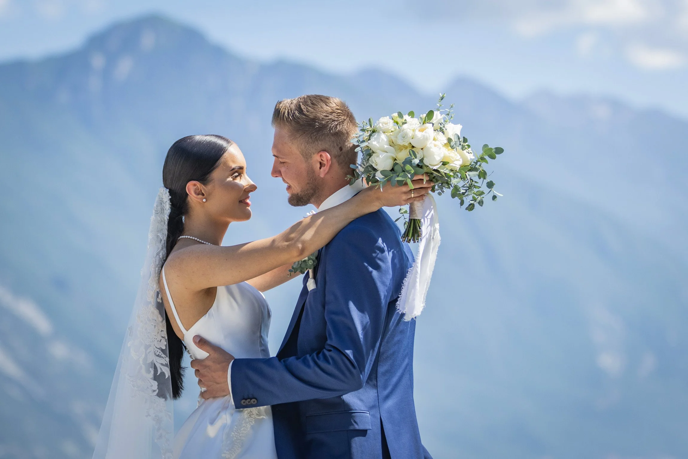Riva del Garda, Trentino. Una coppia di sposi si guarda amorevolmente in un paesaggio montano, la donna indossa un abito bianco con velo e collana di perle, lui un completo blu, la donna tiene un bouquet di fiori bianchi

© Alessandro Holneider
