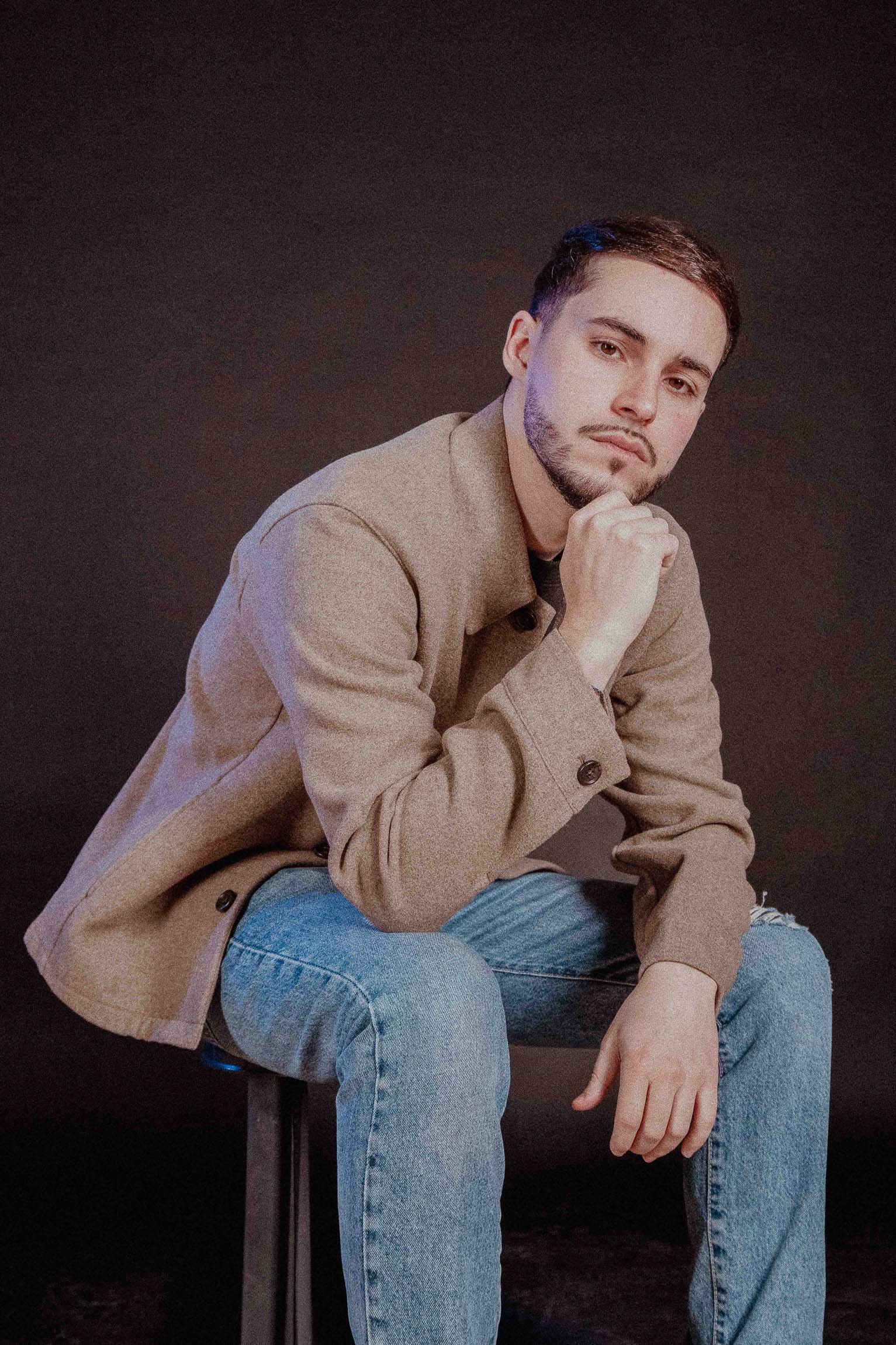 Ragazzo seduto su una sedia, con giacca beige e jeans, posa con il braccio appoggiato sul ginocchio e la mano sotto il mento, sfondo nero.

© Alessandro Holneider