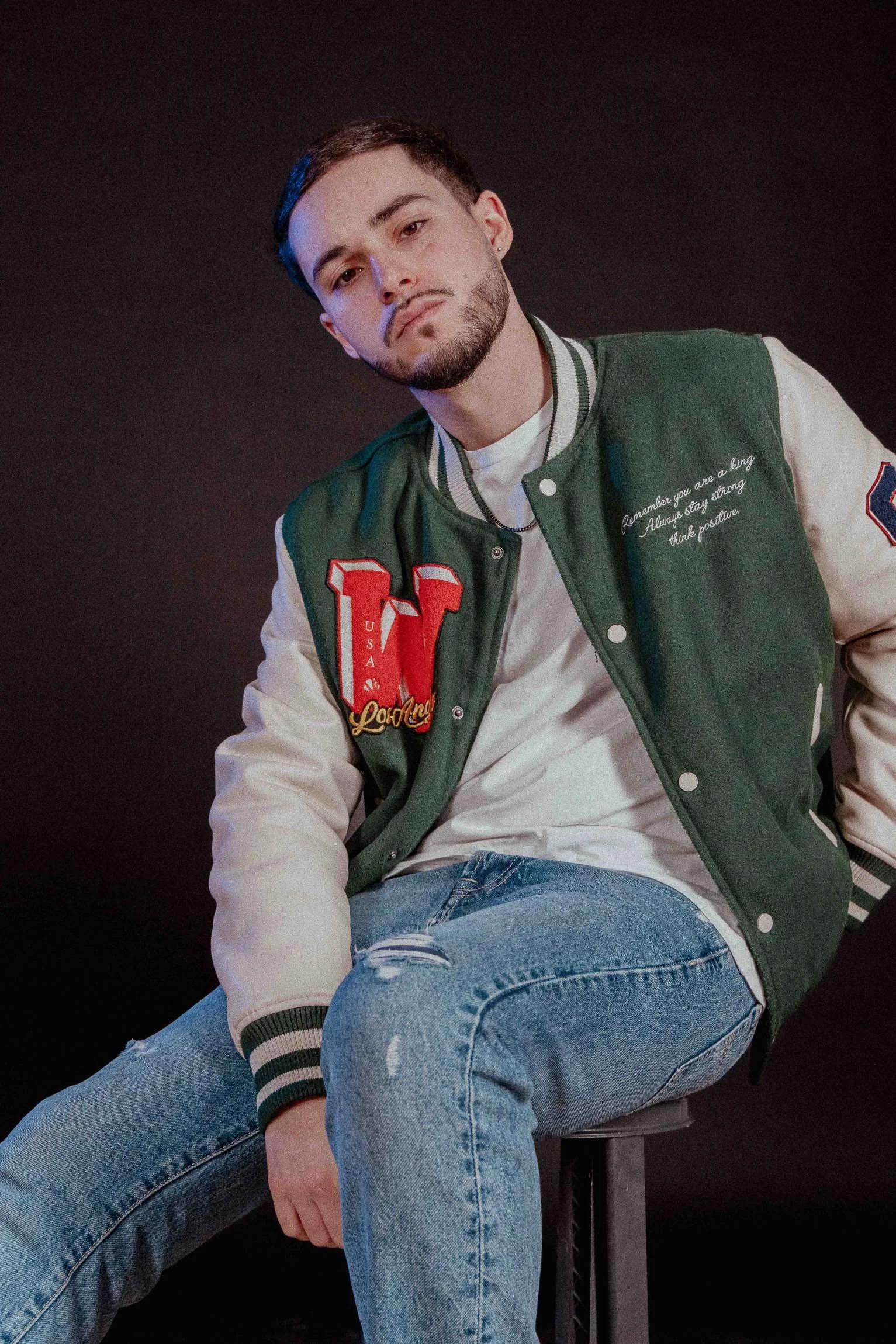 Ragazzo seduto su uno sgabello, indossa una giacca varsity verde con dettagli bianchi e pantaloni jeans strappati.

© Alessandro Holneider