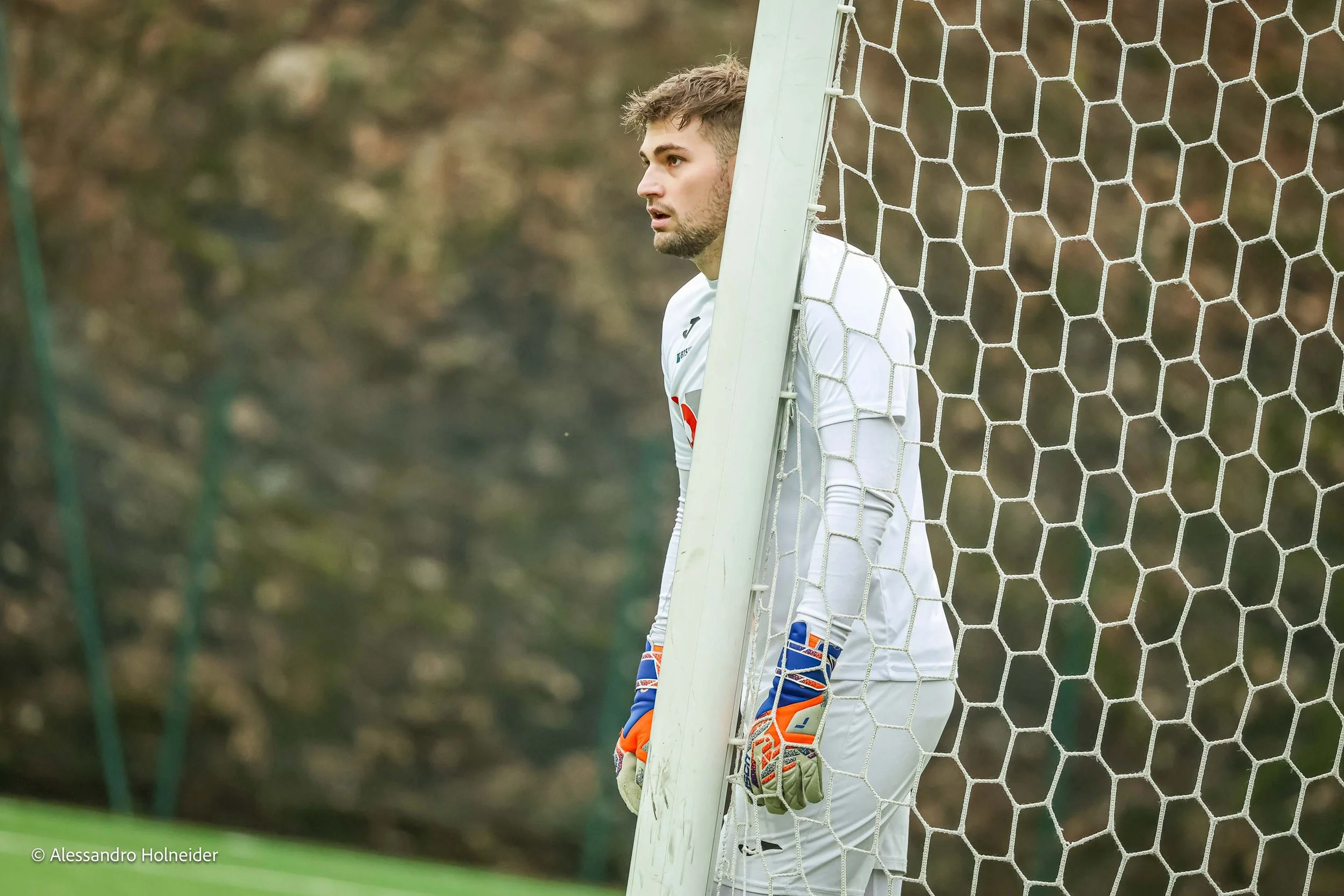 Portiere di calcio in divisa bianca con guanti arancioni e blu, in piedi dietro la porta su un campo di calcio, con un'espressione concentrata.