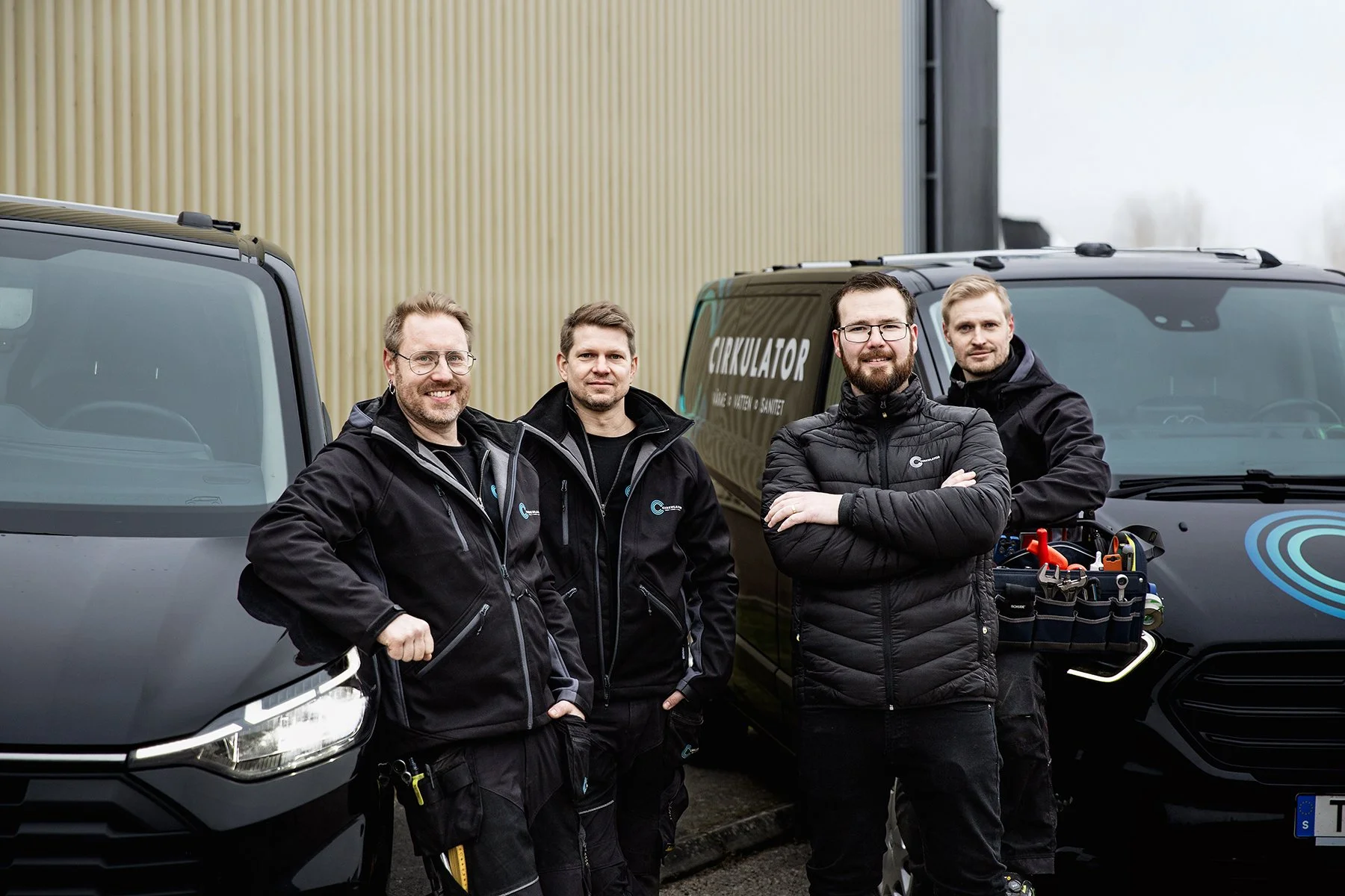 Fyra män står utomhus framför två svarta fordon, alla bär svarta jackor och ser glada ut.