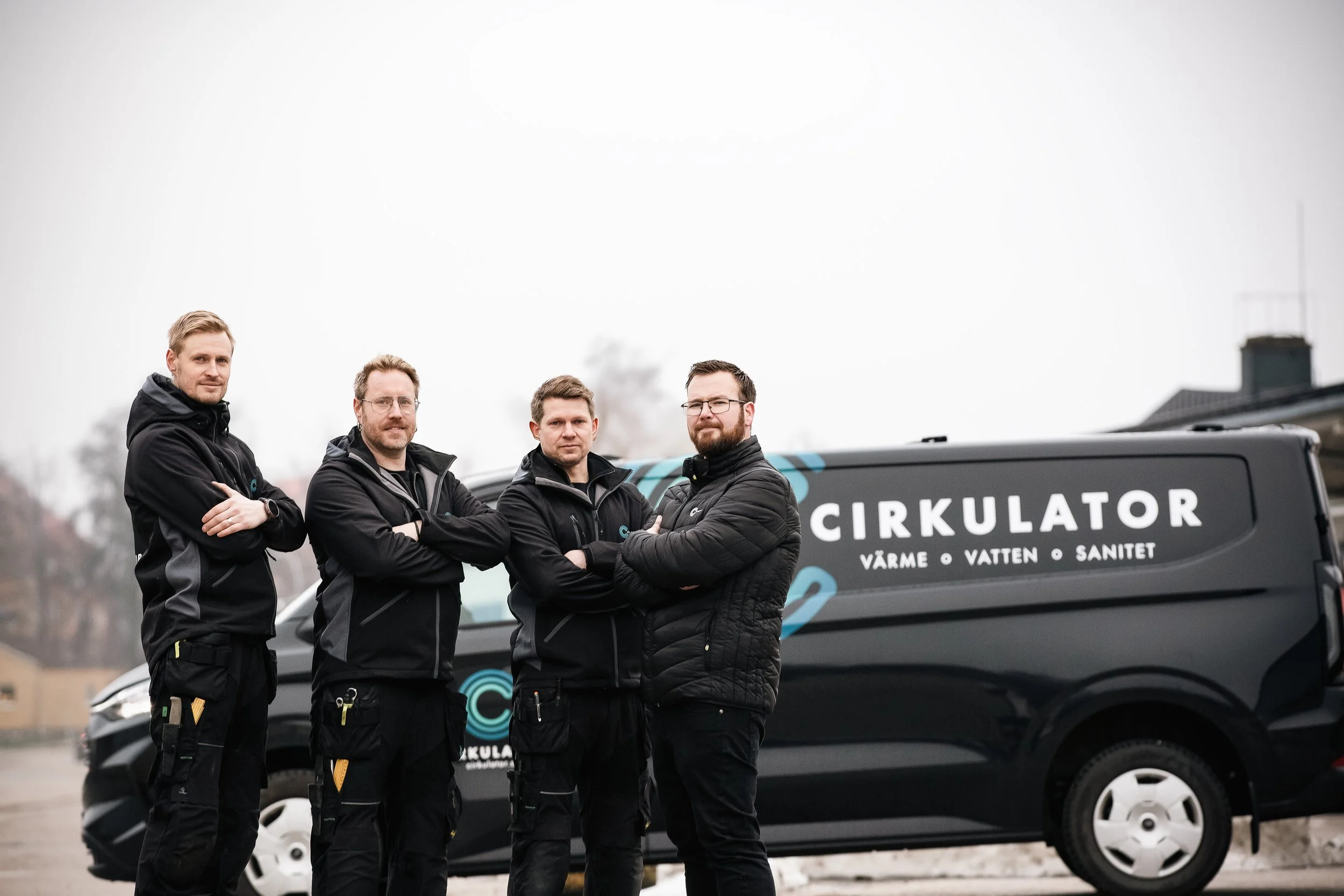 Fyra män står med korsade armar framför en svart tjänstebil med texten 'Cirkulator Värme Vatten Sanitet'.