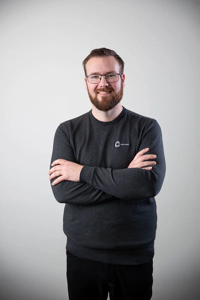 VD Andreas med glasögon och skägg, bär en mörkgrå tröja, står fram emot en vit vägg, ler mot kameran.