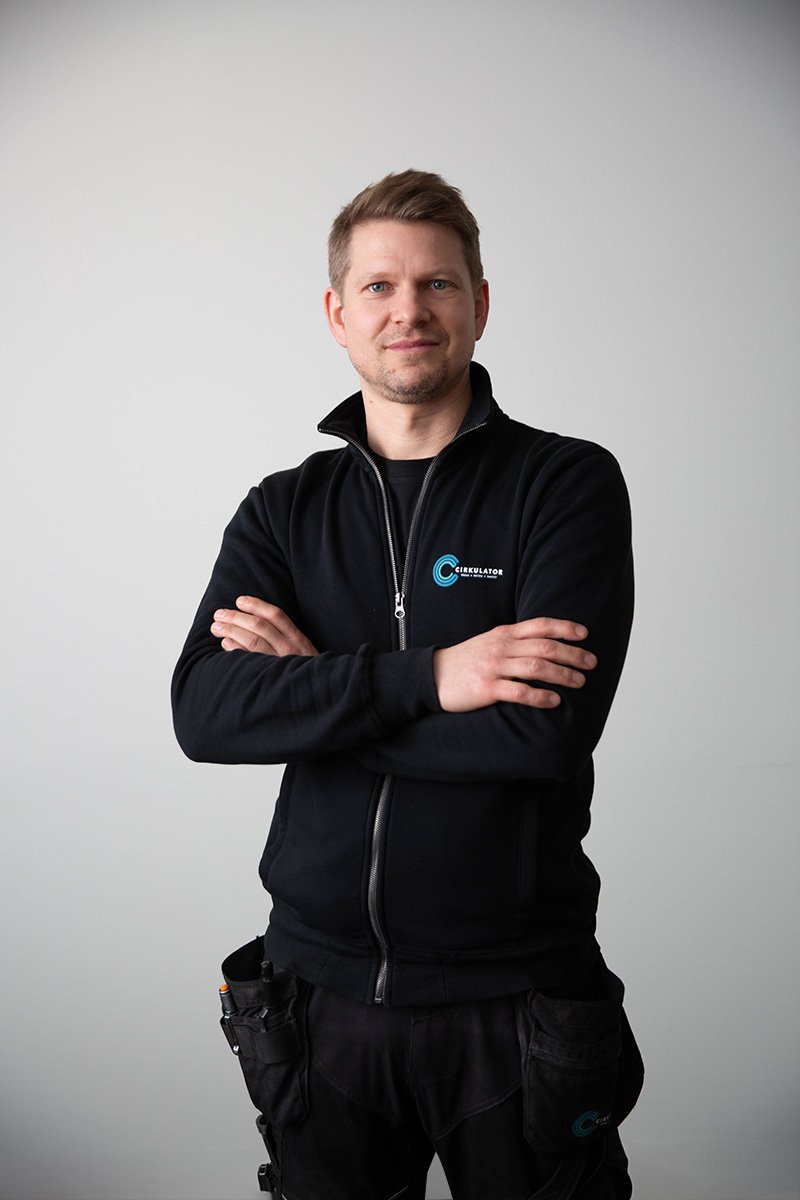 Man står mot en vit vägg, klädd i en svart jacka med logga på bröstet, med armarna korsade.