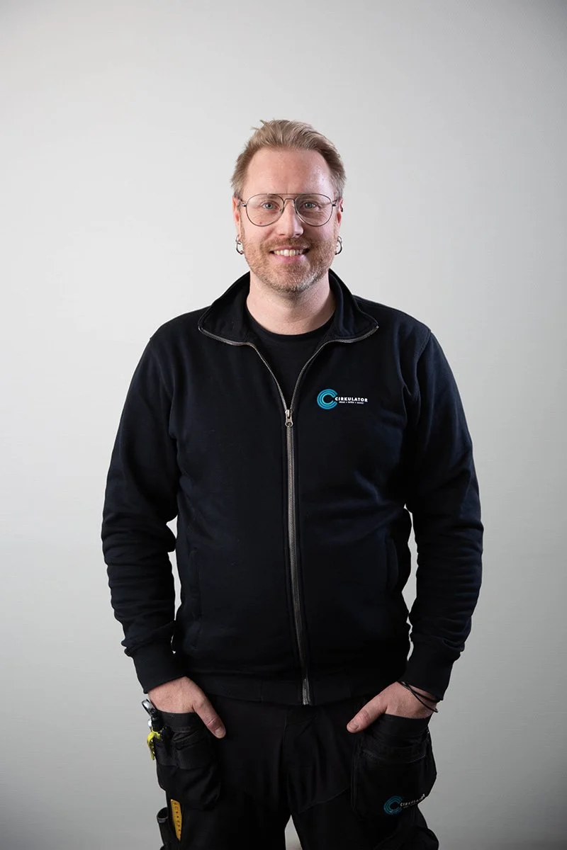 Kollega med läsglasögon, skägg och örhängen, bär svart jacka med logga och står framför en vit vägg.