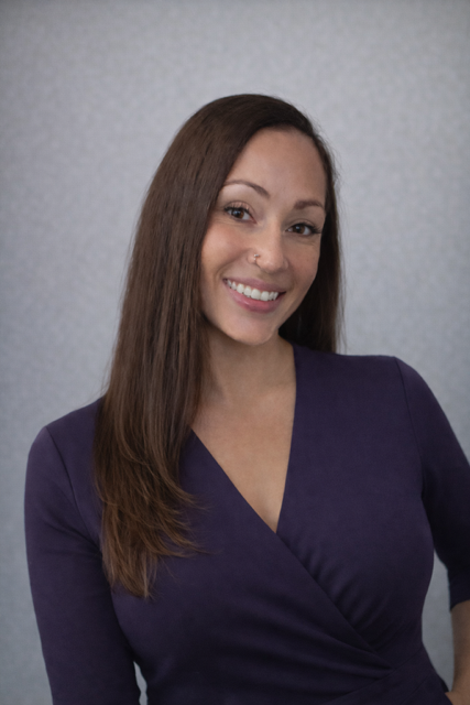A woman with long brown hair, smiling, wearing a purple V-neck top with a textured gray background.