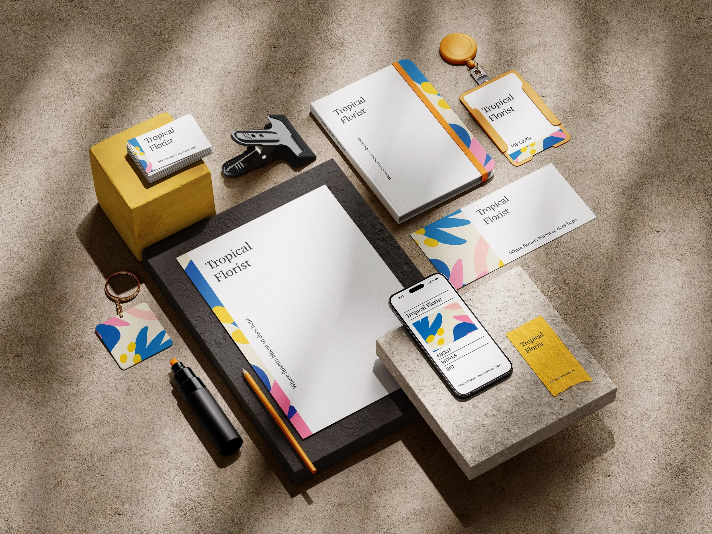 Flat lay of business branding materials and office supplies for Tropical Florist, including business cards, a notebook, a smartphone with the company website, a keychain, a pencil, a small bottle, a badge reel, and several paper sheets with colorful abstract designs.