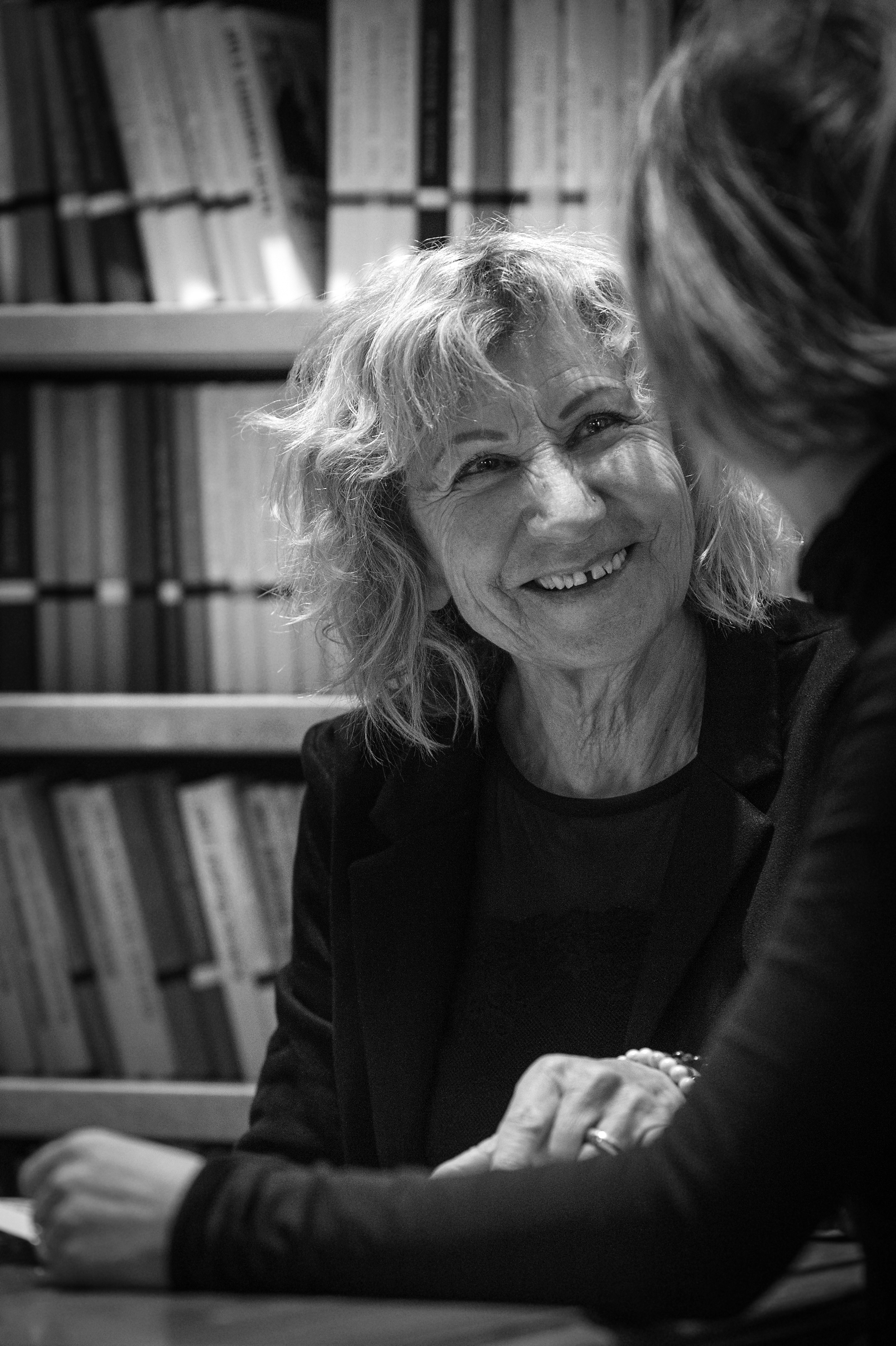 Black and white photo of two women talking, one smiling warmly, in front of a bookshelf filled with books.