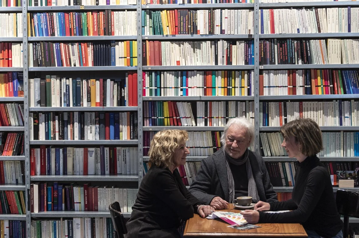 Three people, two women and one man, sitting at a wooden table and having a conversation in a library with tall bookshelves filled with colorful books in the background.