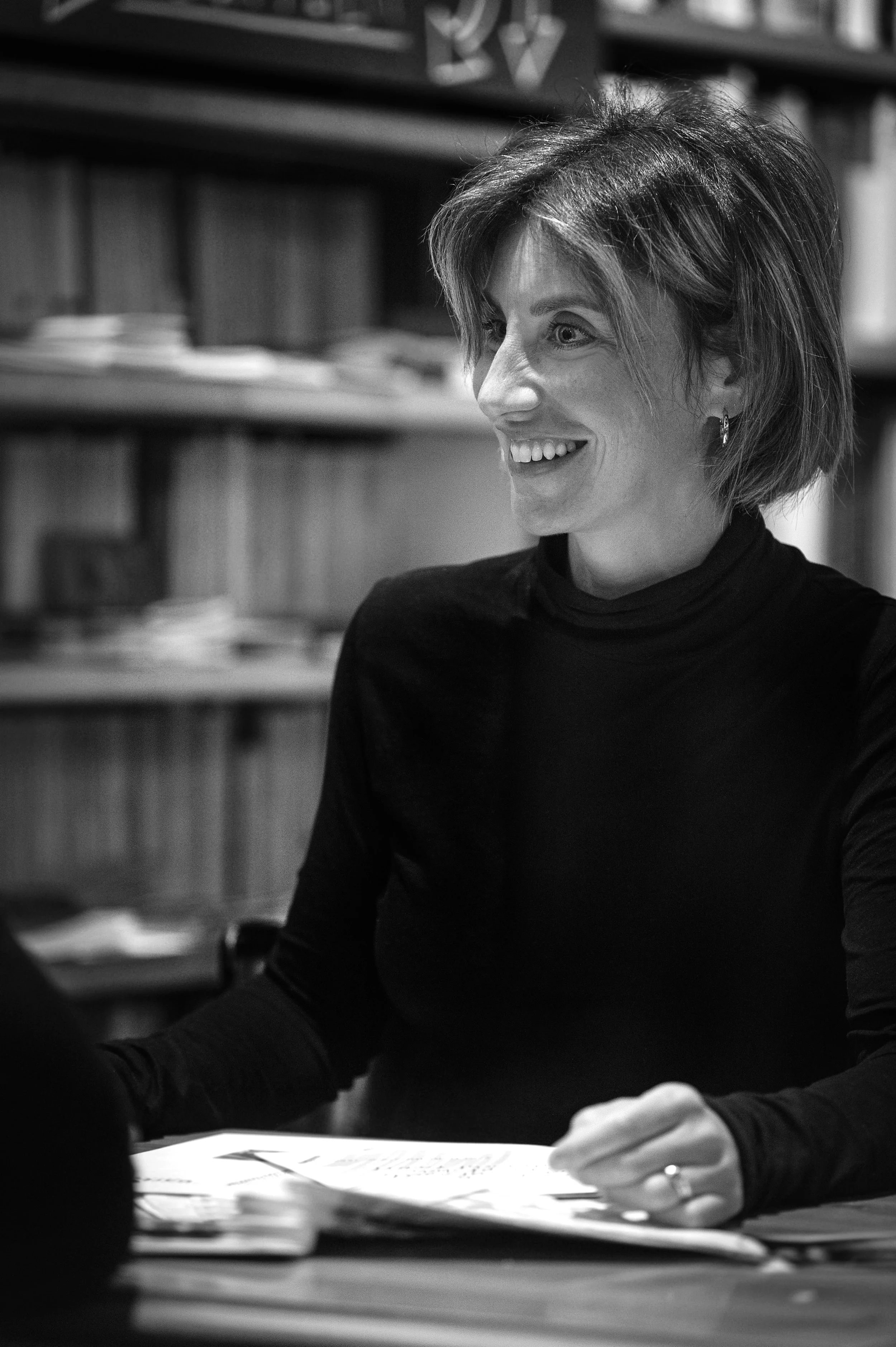 Black and white photo of a woman with short hair, smiling and sitting at a table in an office or library with bookshelves in the background. Seraina Appiah-Alder