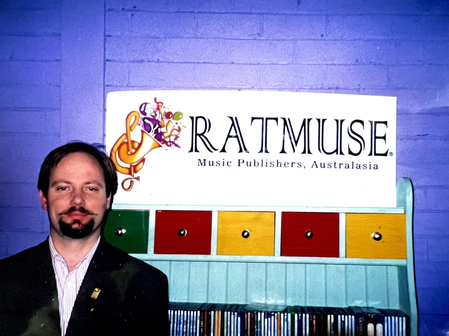 A man with dark hair and a beard, wearing a dark suit and a white shirt, standing in front of a sign that reads 'RATMUSE Music Publishers, Australasia.' The sign is mounted on a textured blue wall, and there is a colorful abstract logo to the left of the text, featuring musical instruments and notes. Below the sign, there is a colorful small cabinet with four drawers in green, red, yellow, and red again, with black knobs.