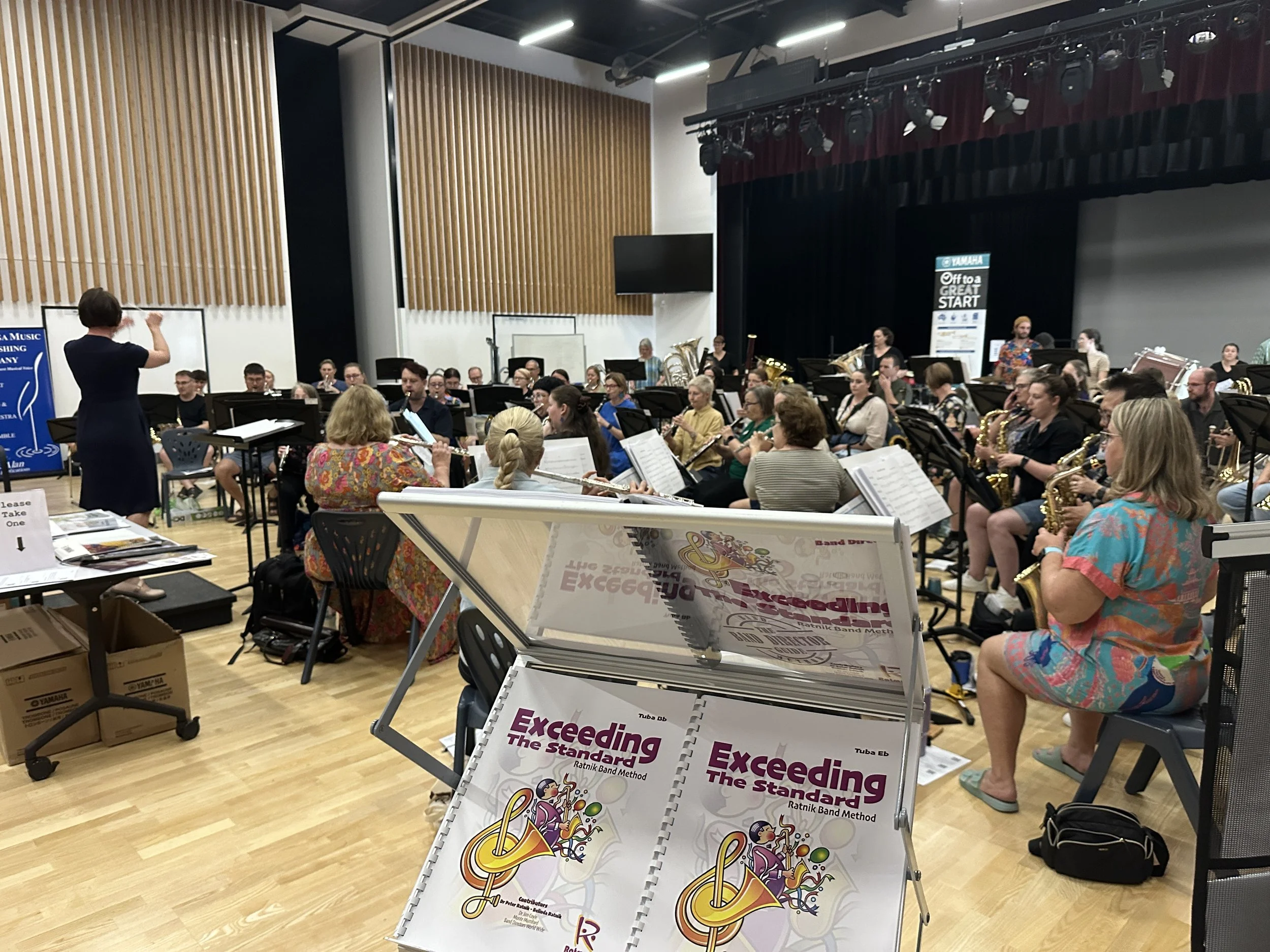 A large group of musicians seated in an auditorium, with a conductor standing in front of them, conducting. The ensemble is a Concert Band. In the foreground the Ratmuse exhibition booth can be seen displaying Exceeding the Standard Band Method.