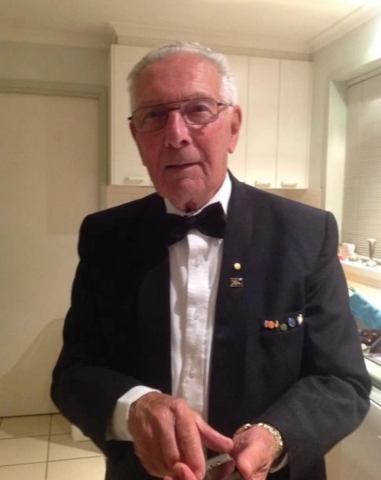 An elderly man in a tuxedo and bow tie, wearing glasses, standing indoors in front of a kitchen. He has a pin and ribbons on his jacket and is clasping his hands.