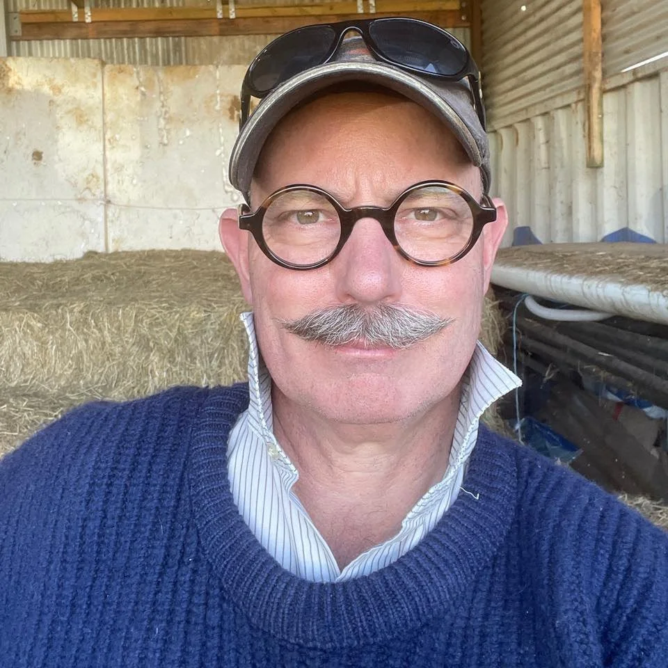Dr Peter Ratnik - A man with a large mustache, glasses, and a cap takes a selfie in a barn or shed with hay and tools in the background.