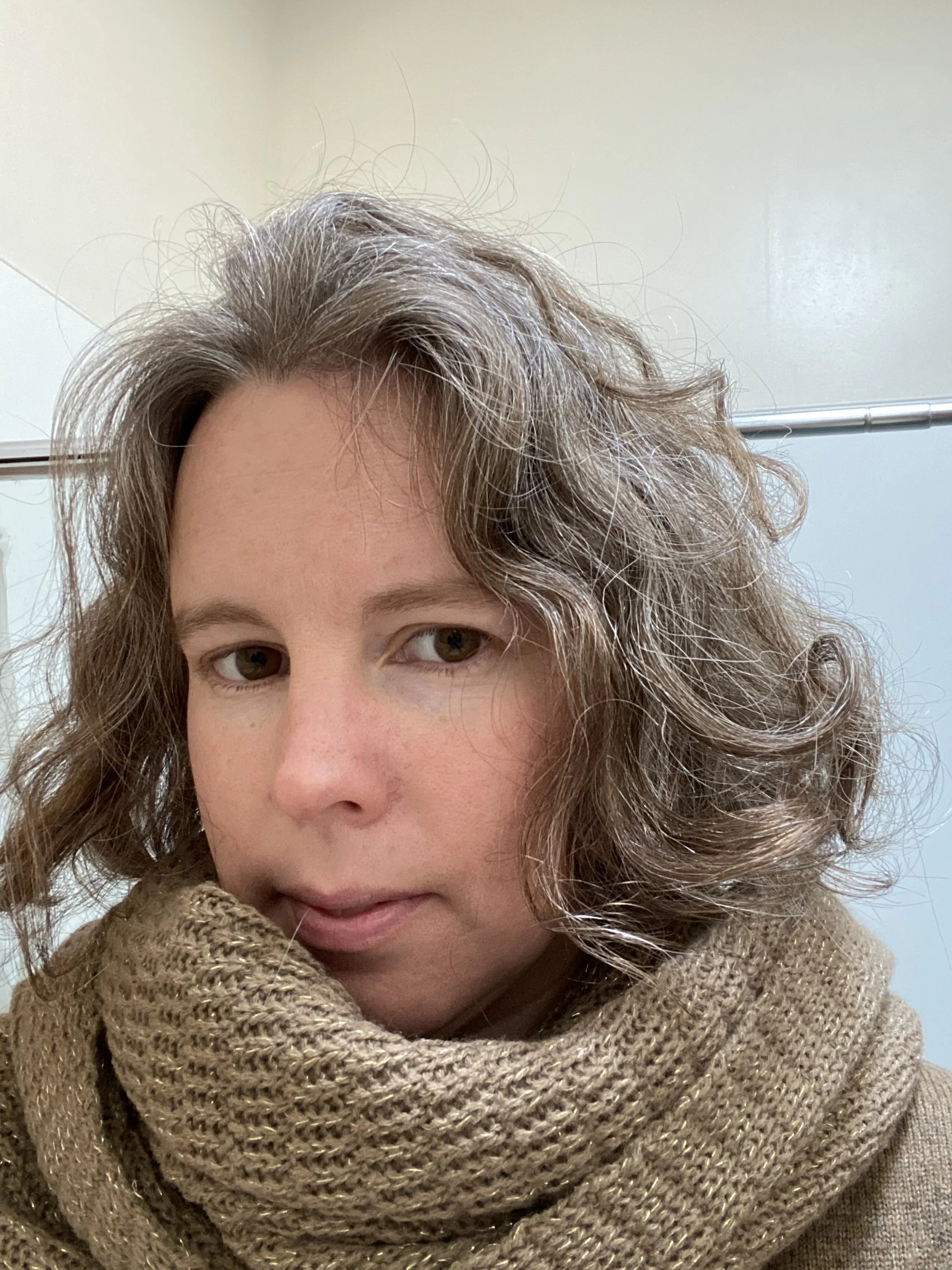 Belinda Ratnik - Close-up of a woman's face with curly hair, wearing a thick, knitted beige scarf.