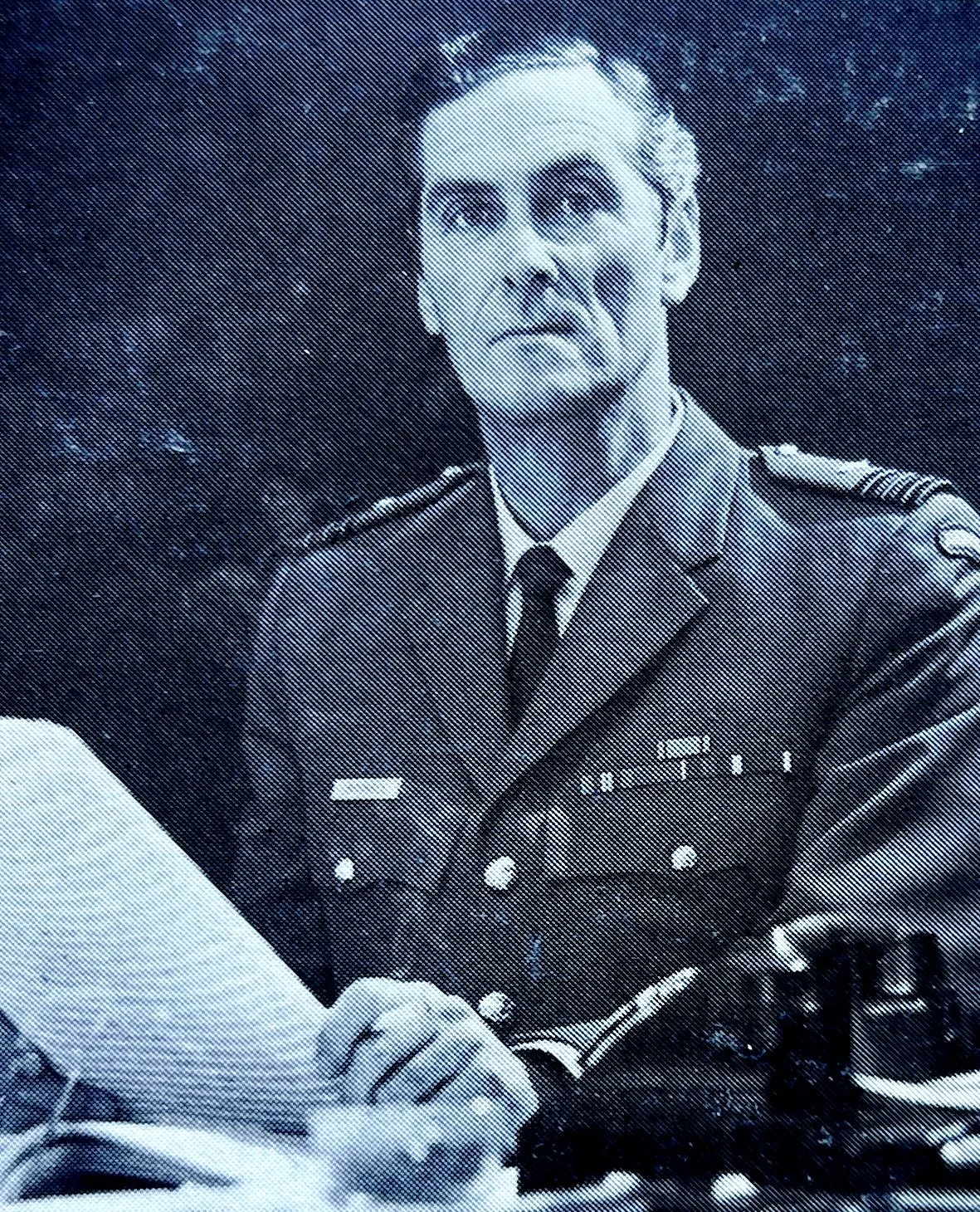 Black and white photo of a man in a military uniform with medals, sitting at a desk and looking slightly upward with a serious expression.