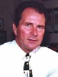 Portrait of a man wearing a white shirt and a patterned tie, with dark hair and a serious expression, in an indoor setting.