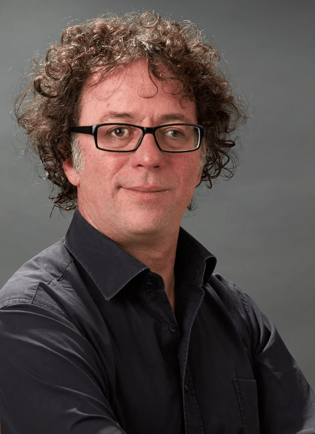 A man with curly gray hair and glasses is wearing a black shirt and looking at the camera with a slight smile, standing against a gray background.
