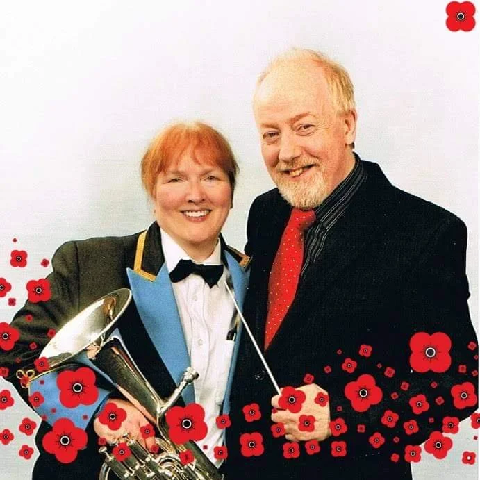 Two men, one in a tuxedo with a blue lapel and bow tie, holding a trophy, and the other in a black suit with a red tie, smiling in front of a white background with red flowers overlay.