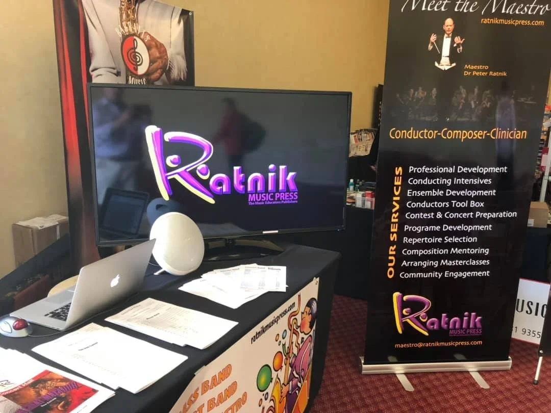 Exhibition booth for Ratnik Music Press displaying a laptop, papers, and a monitor screen, with a black vertical banner listing music-related services and a poster of a conductor.
