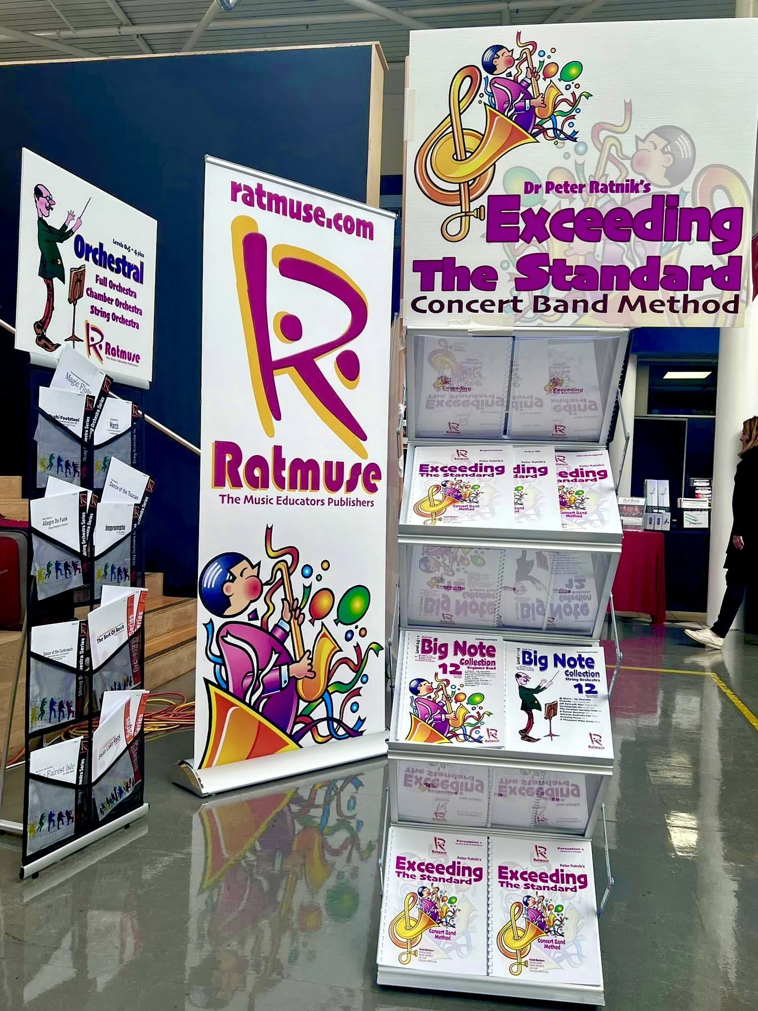 Display booth for music educational publisher, featuring books and posters about Dr. Peter Ratnik's Exceeding The Standard Concert Band Method, with colorful cartoon illustrations of a musician playing a saxophone and a conductor.