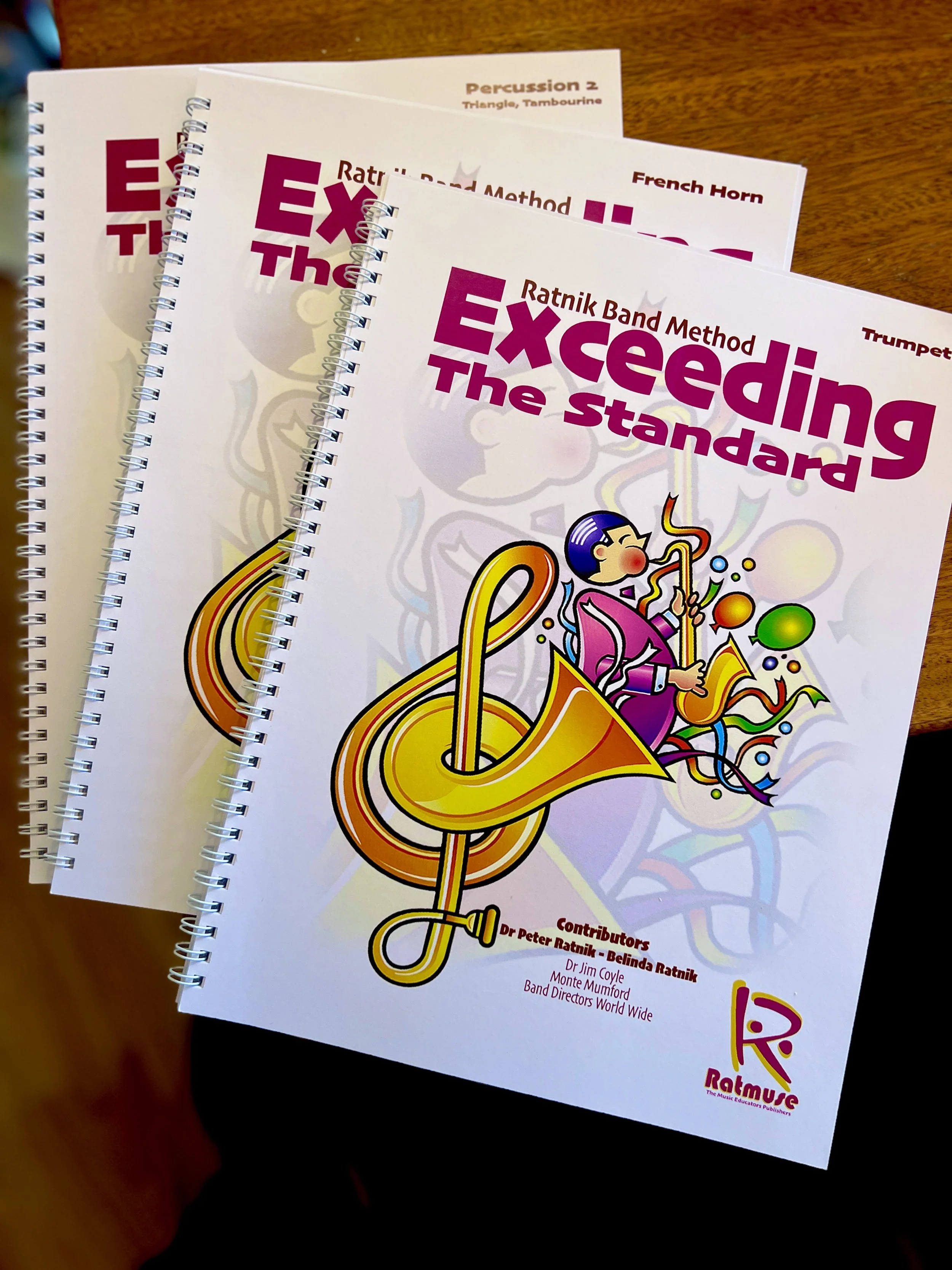 Multiple copies of a colorful music educational book titled 'Exceeding the Standard' with illustrations of musical instruments and characters, placed on a wooden surface.