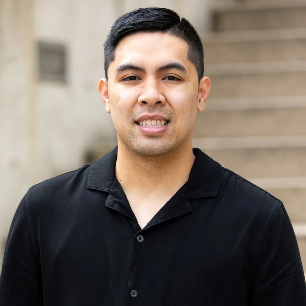 A young man with short black hair, wearing a black collared shirt, standing outdoors in front of a staircase.