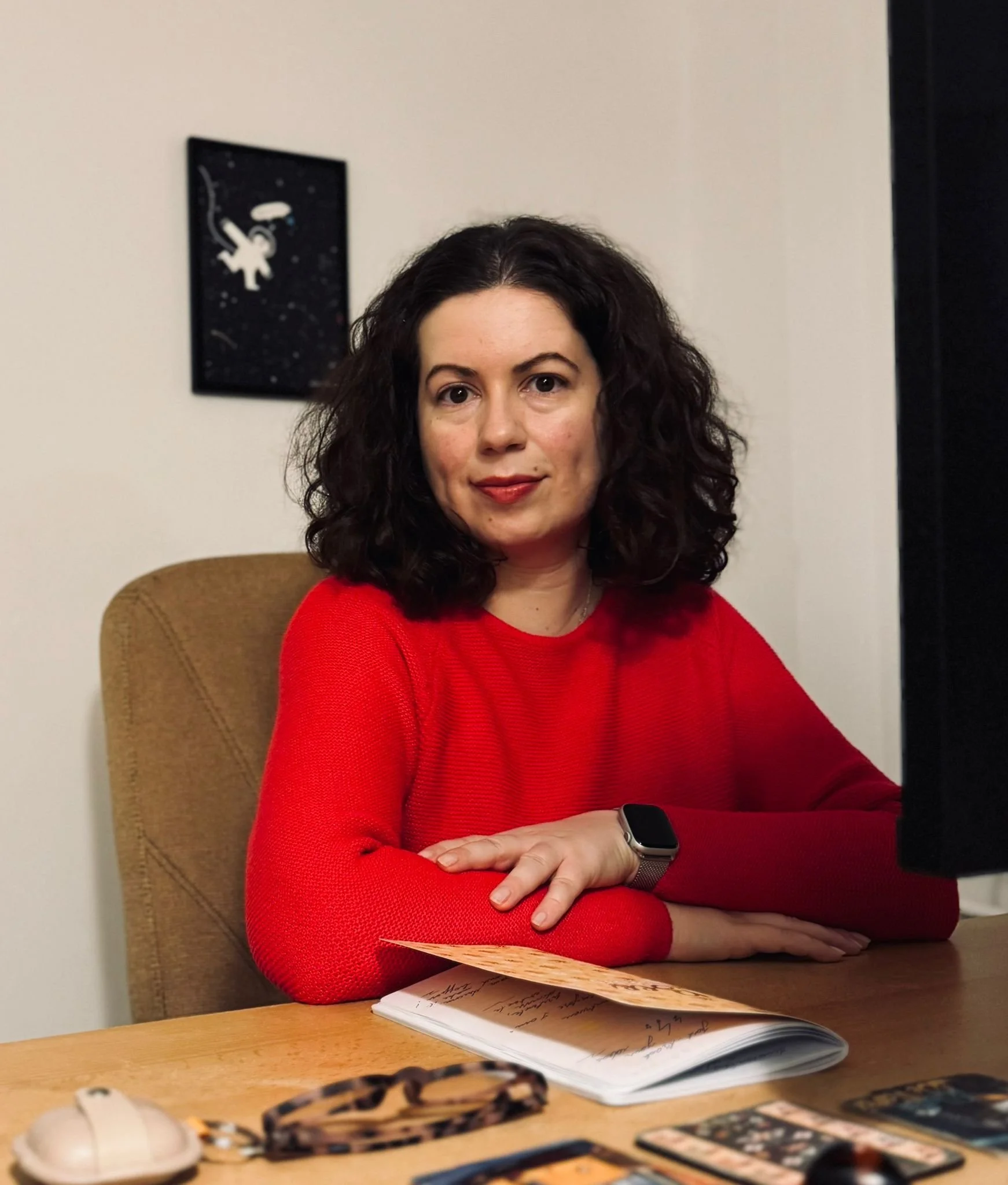 A woman with dark curly hair and a red sweater sitting at a desk with a notebook and miscellaneous items, looking at the camera.