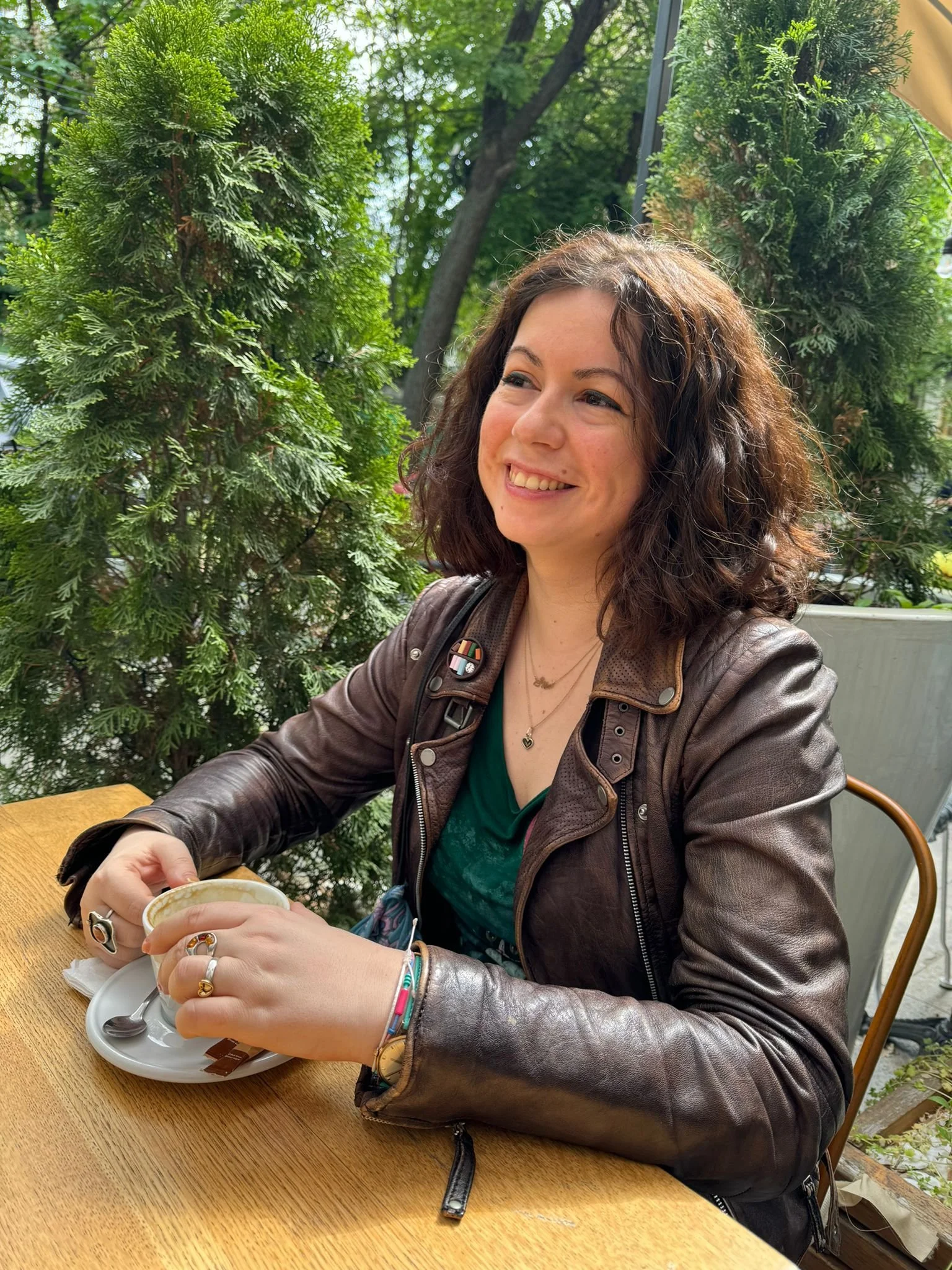 A woman with curly brown hair smiling while sitting at a wooden table outdoors, holding a cup of coffee with greenery and trees in the background.