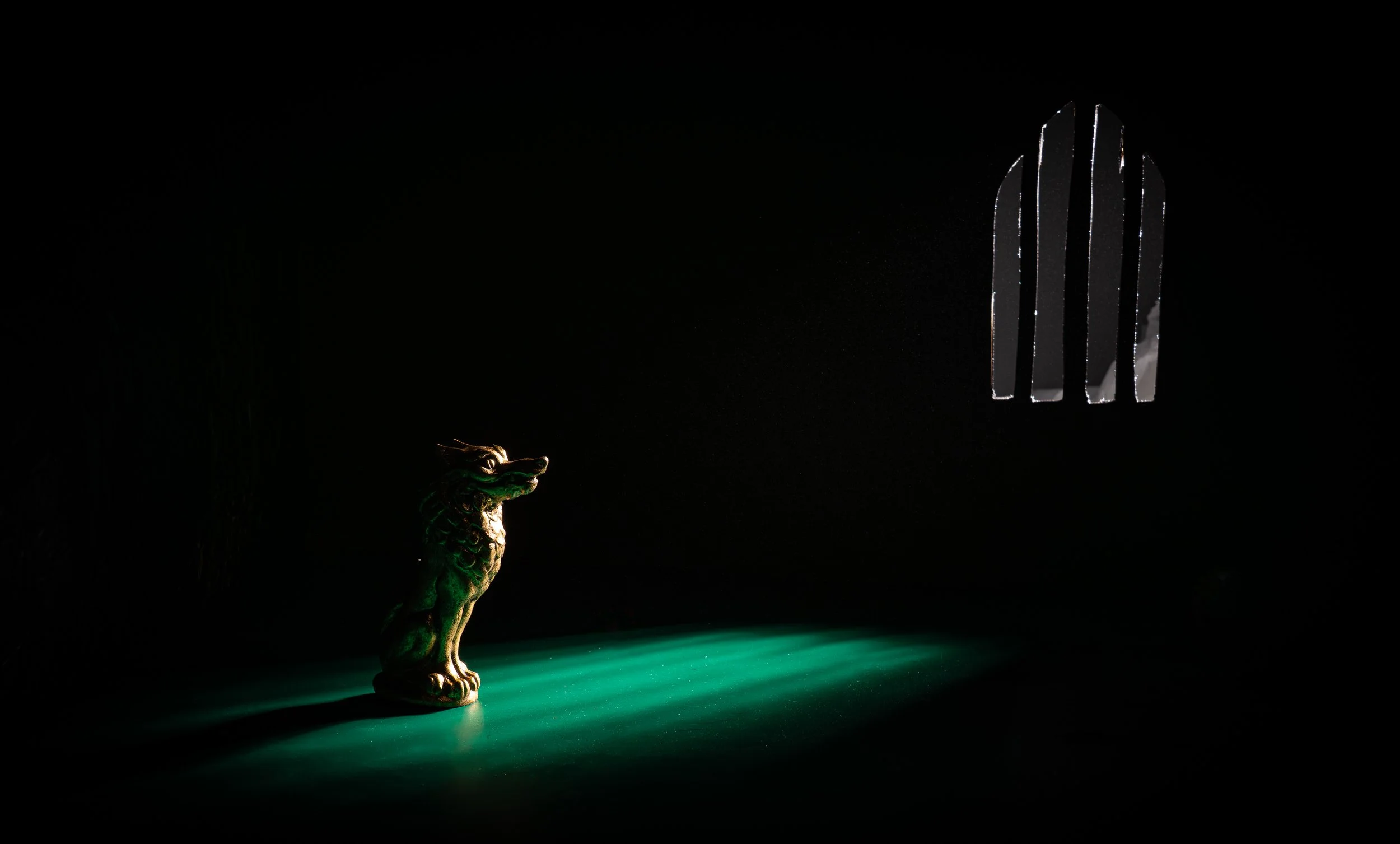 A small, shiny golden lion statue on a dark surface, illuminated by green light, with a barred window in the background.