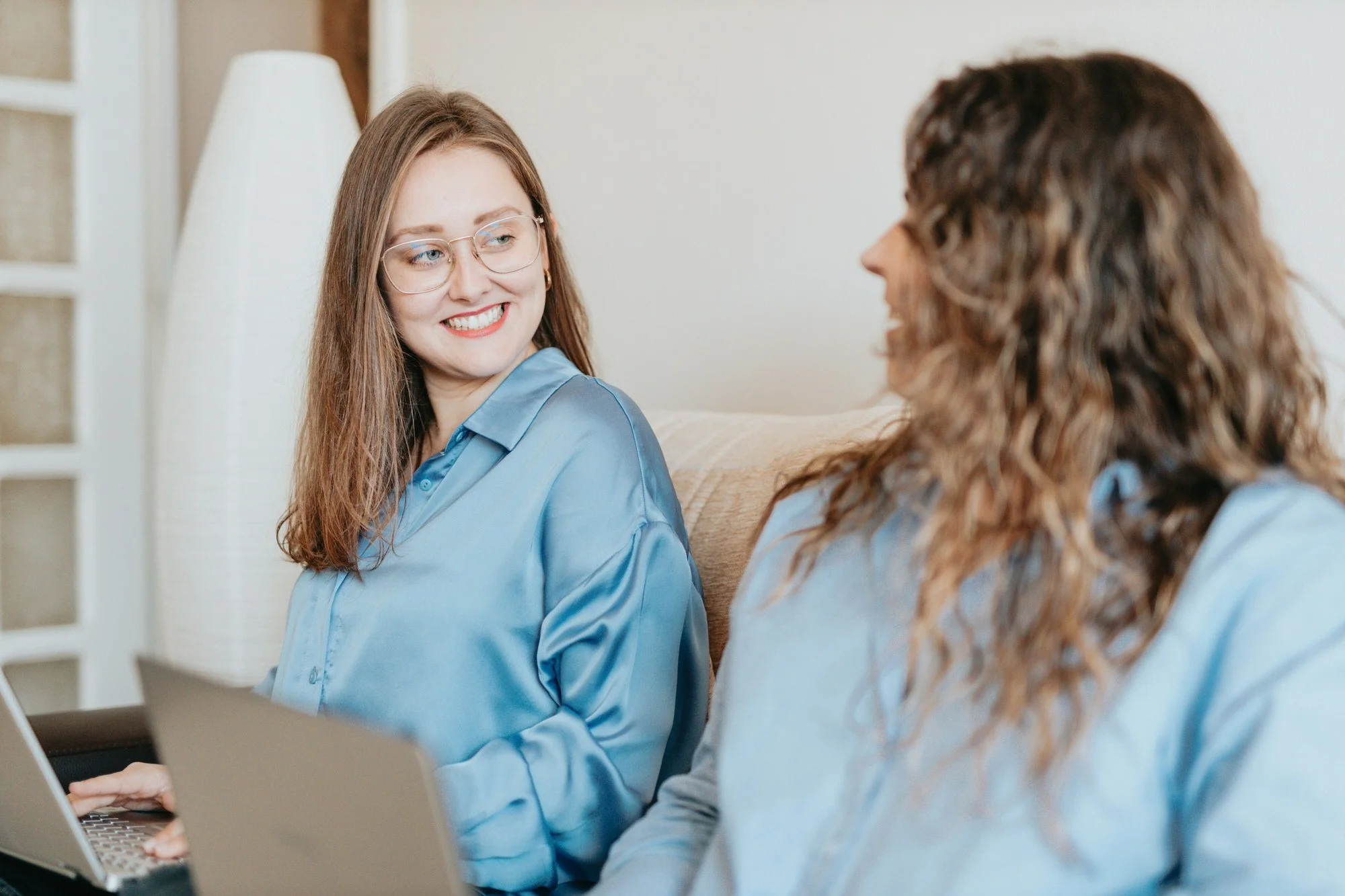 Two women sitting on a beige couch, smiling, facing each other, with laptops open in front of them, wearing blue shirts, in a cozy room with a lamp behind them.