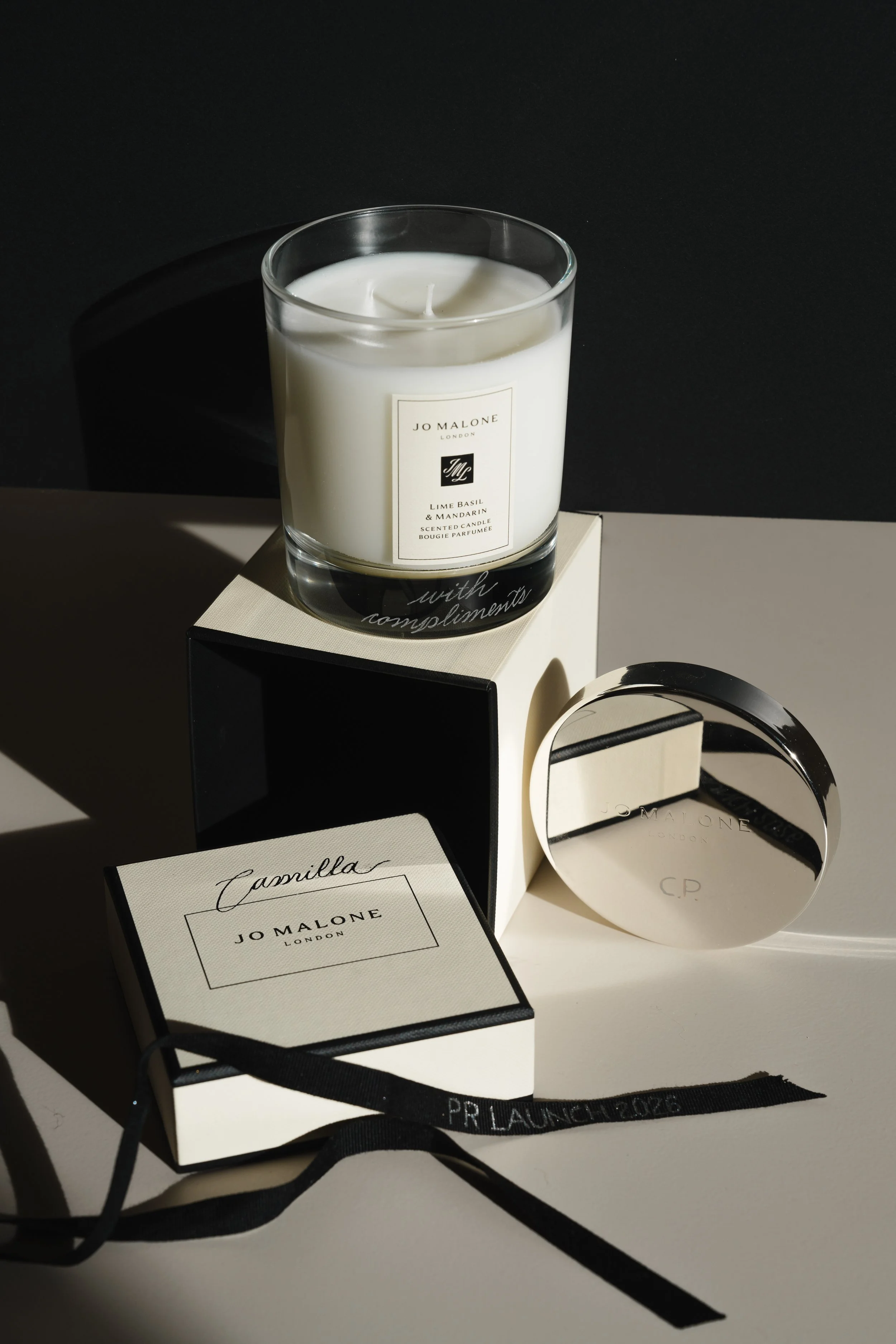 Et håndgravert og personalisert duftlys i glass med merke Jo Malone, plassert på en svart og hvit eske, omgitt av sorte bånd, mot en mørk bakgrunn.