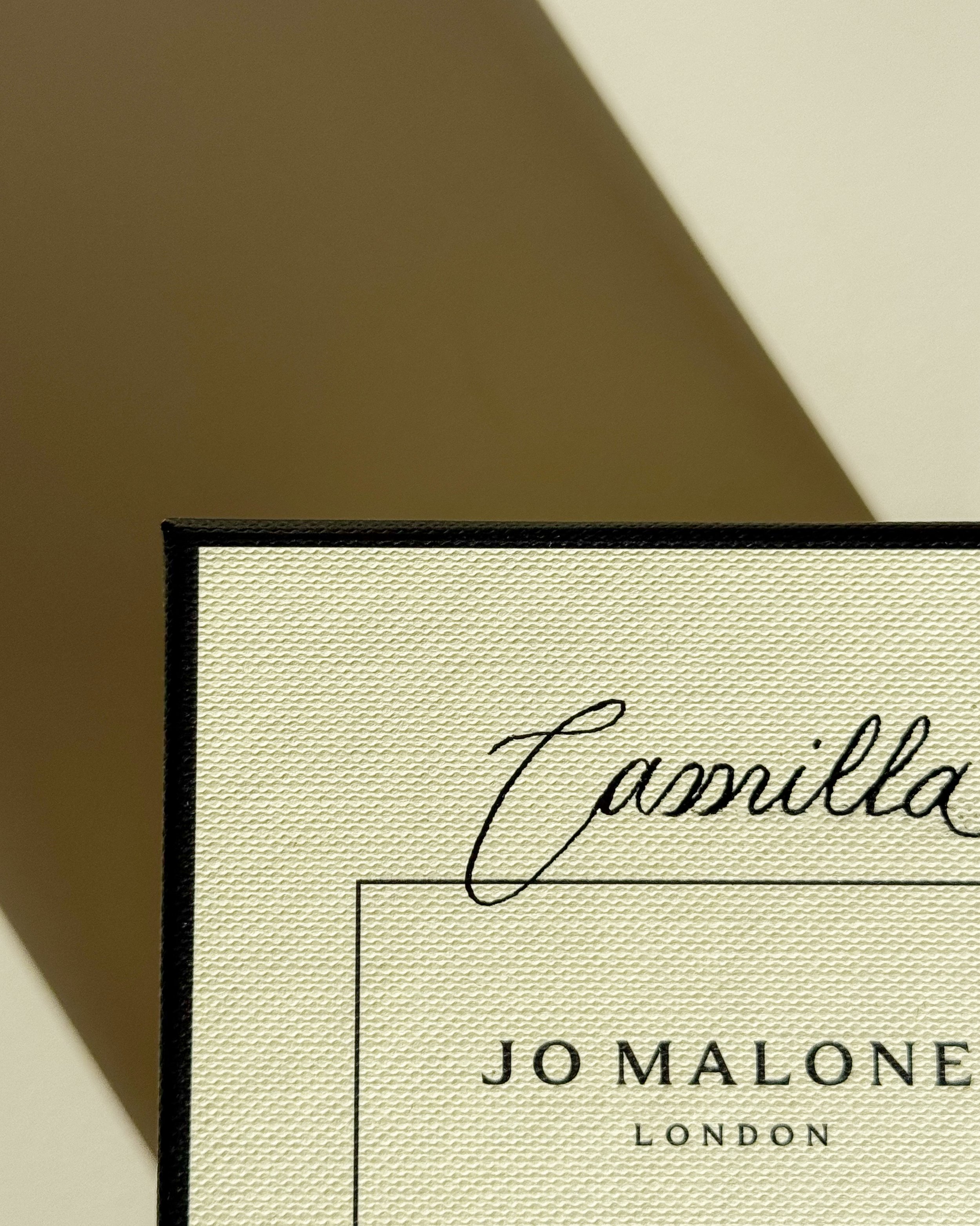 	
Bildet viser en del av en personalisert lukk med navnet "Camilla" og teksten "JO MALONE LONDON". Bakgrunnen er beige, og skiltet har en svart ramme.