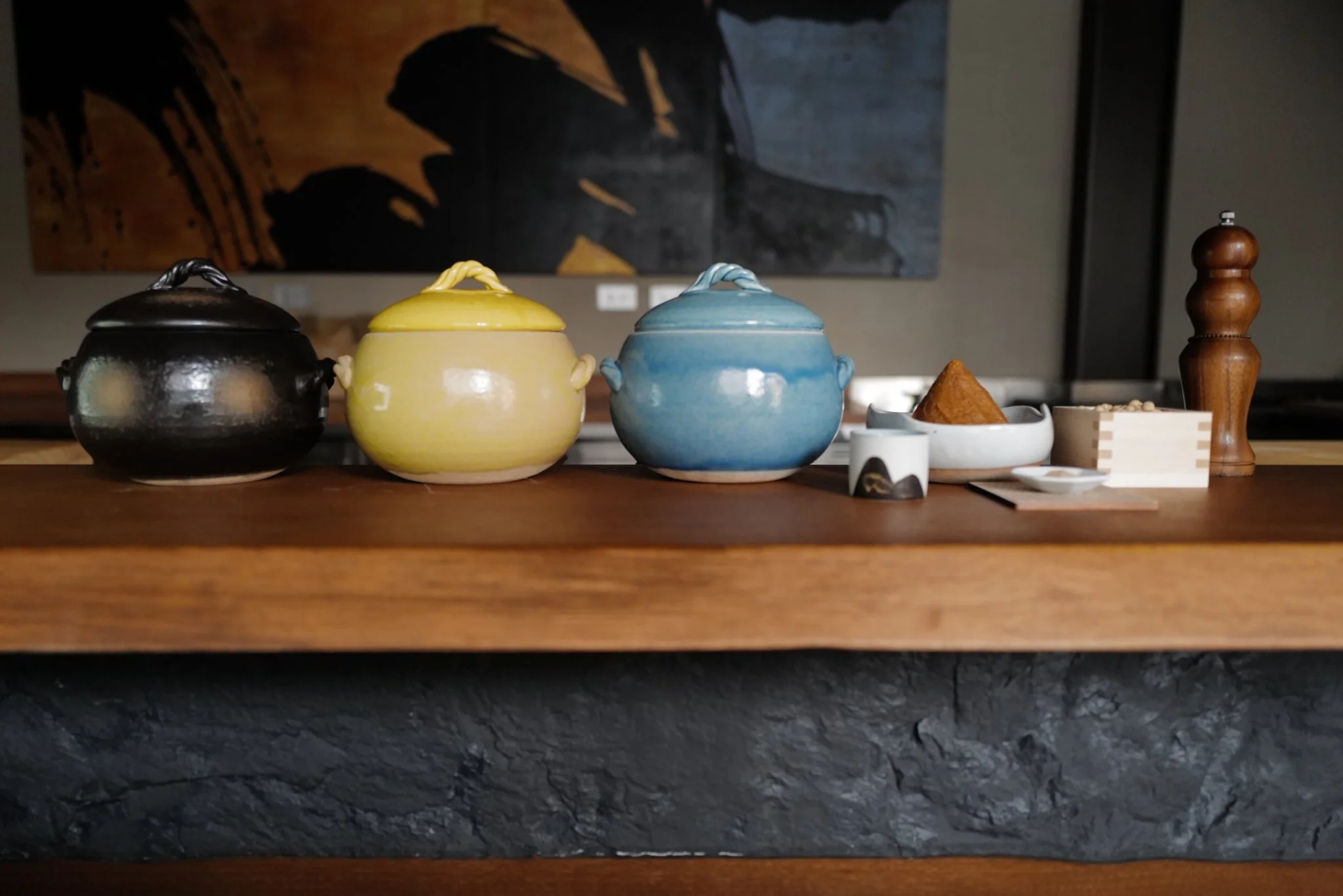 Three colorful ceramic containers with lids in black, yellow, and blue, accompanied by small dishes and a wooden pepper grinder on a wooden surface.