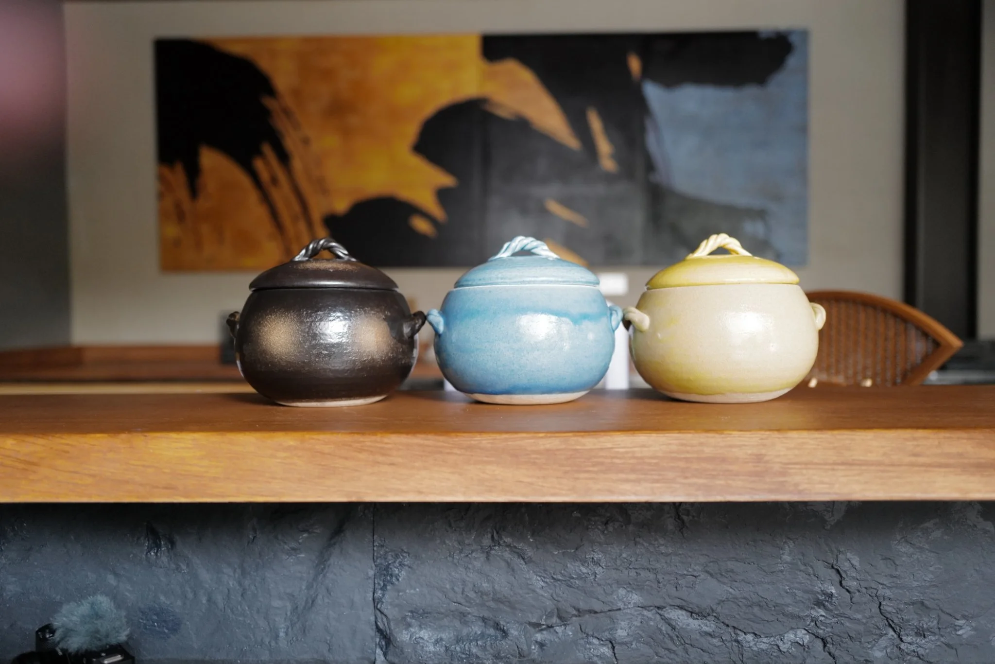 Three ceramic or pottery containers with lids and small handles on a wooden surface, with a painting of a person's face in a warm color palette hanging on the wall behind them.