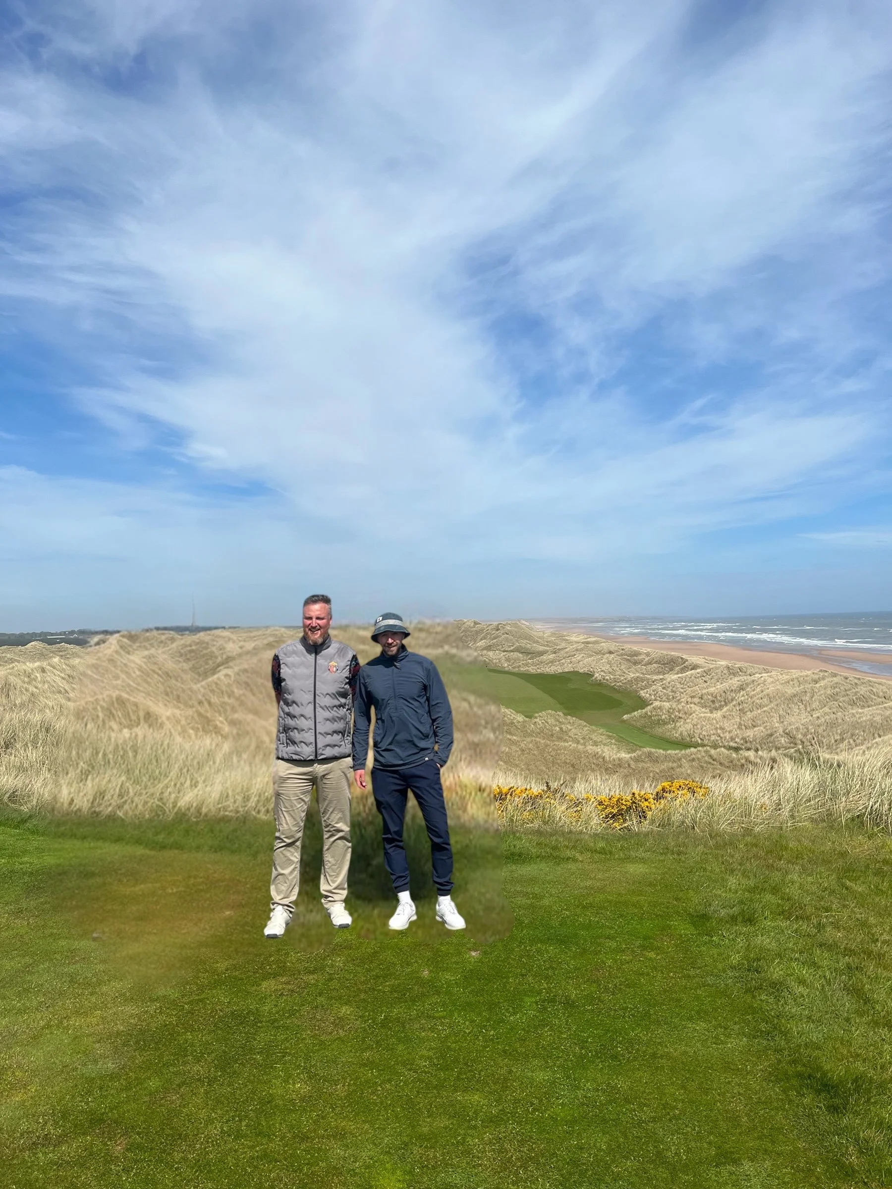 Two men standing on a grassy sand dune landscape with a beach and ocean in the background, under a partly cloudy sky.
