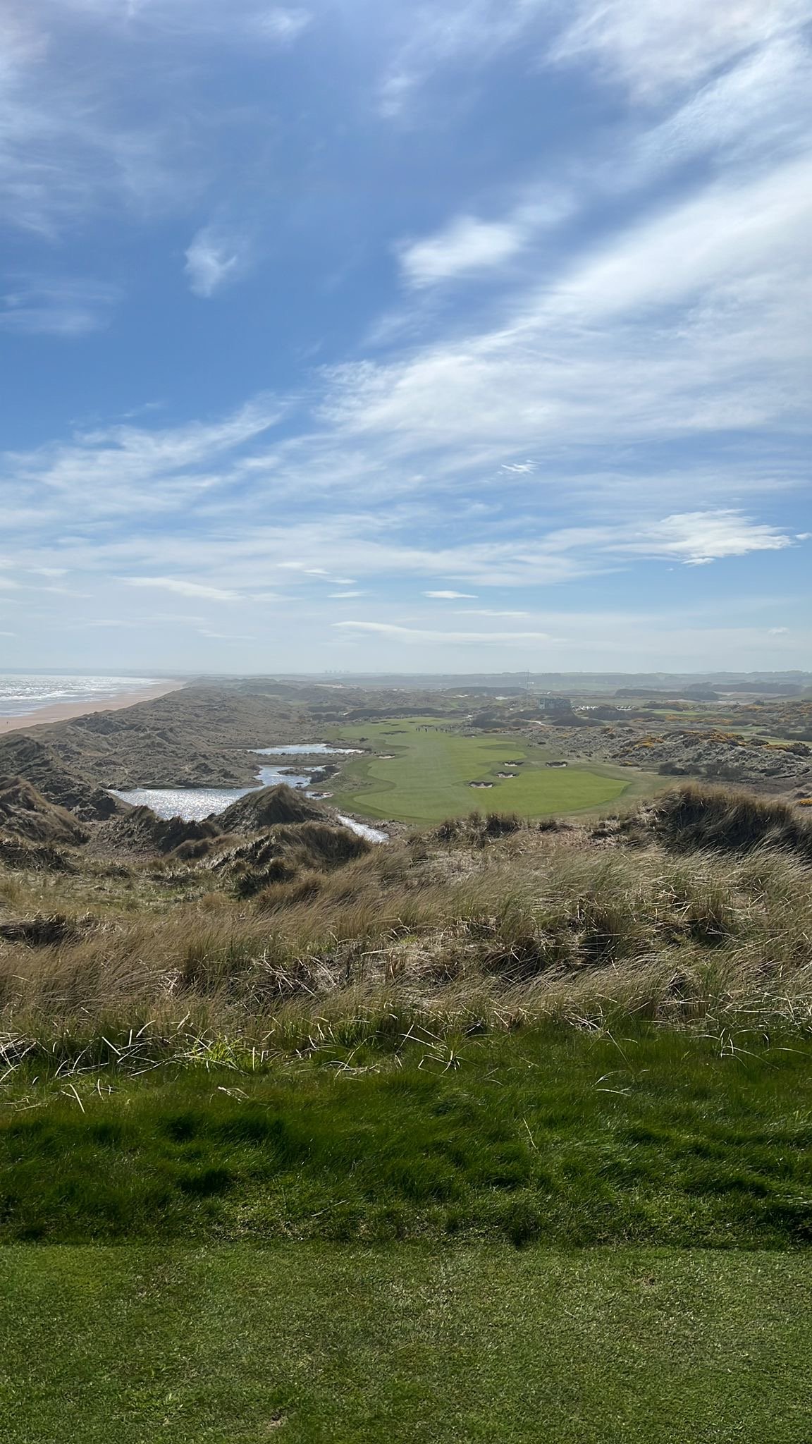 Golf course with grass, dunes, water, and a view of the ocean in the distance under a blue sky with scattered clouds.