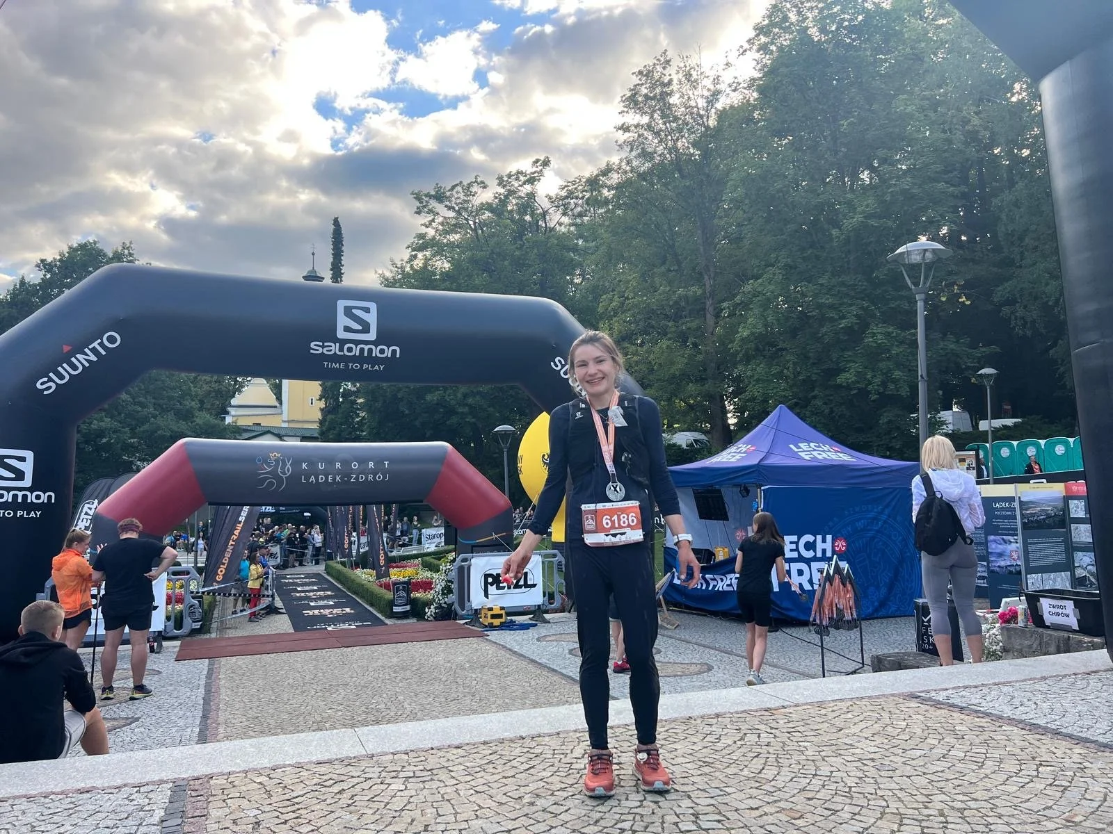 A woman in running gear with a medal on stands at the end of a race finish line, smiling at the camera