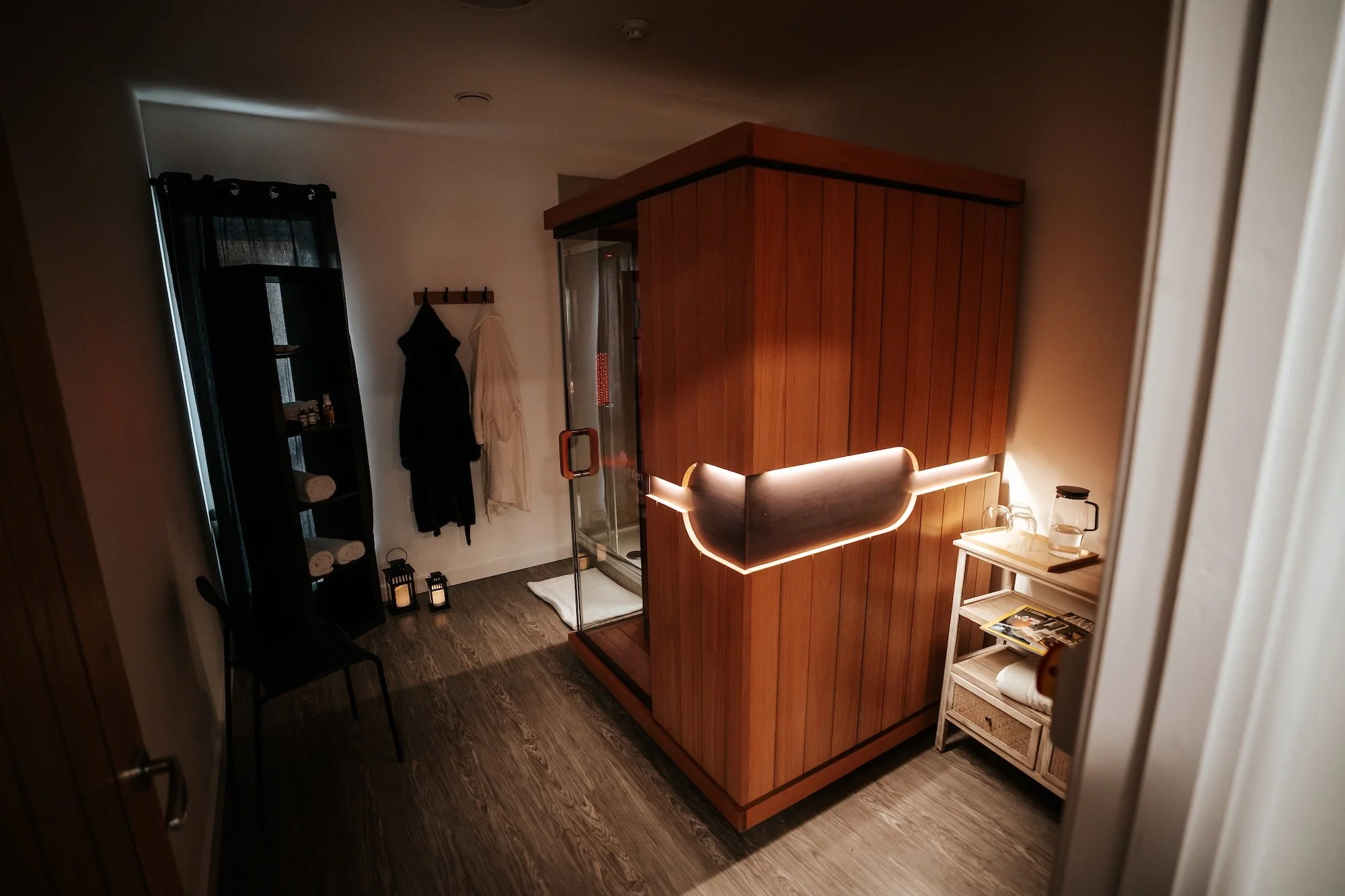 A dimly light room showing an infrared sauna, hydration area, towels and dressing downs hanging up