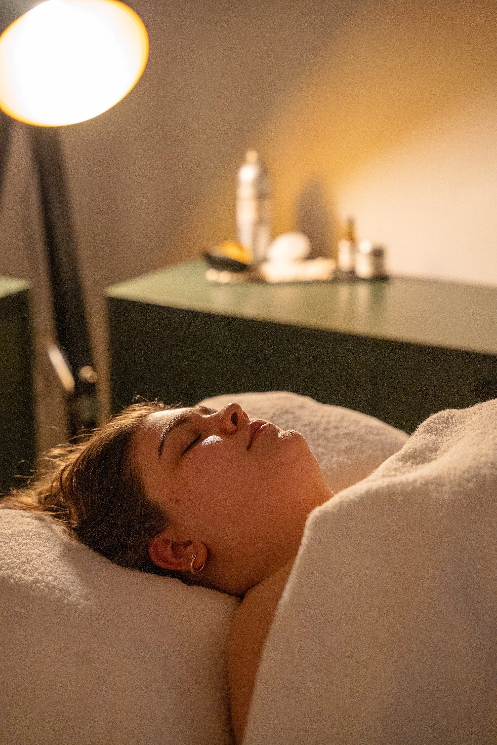 A woman lies with her eyes on closed on a treatment bed, covering in a white towel
