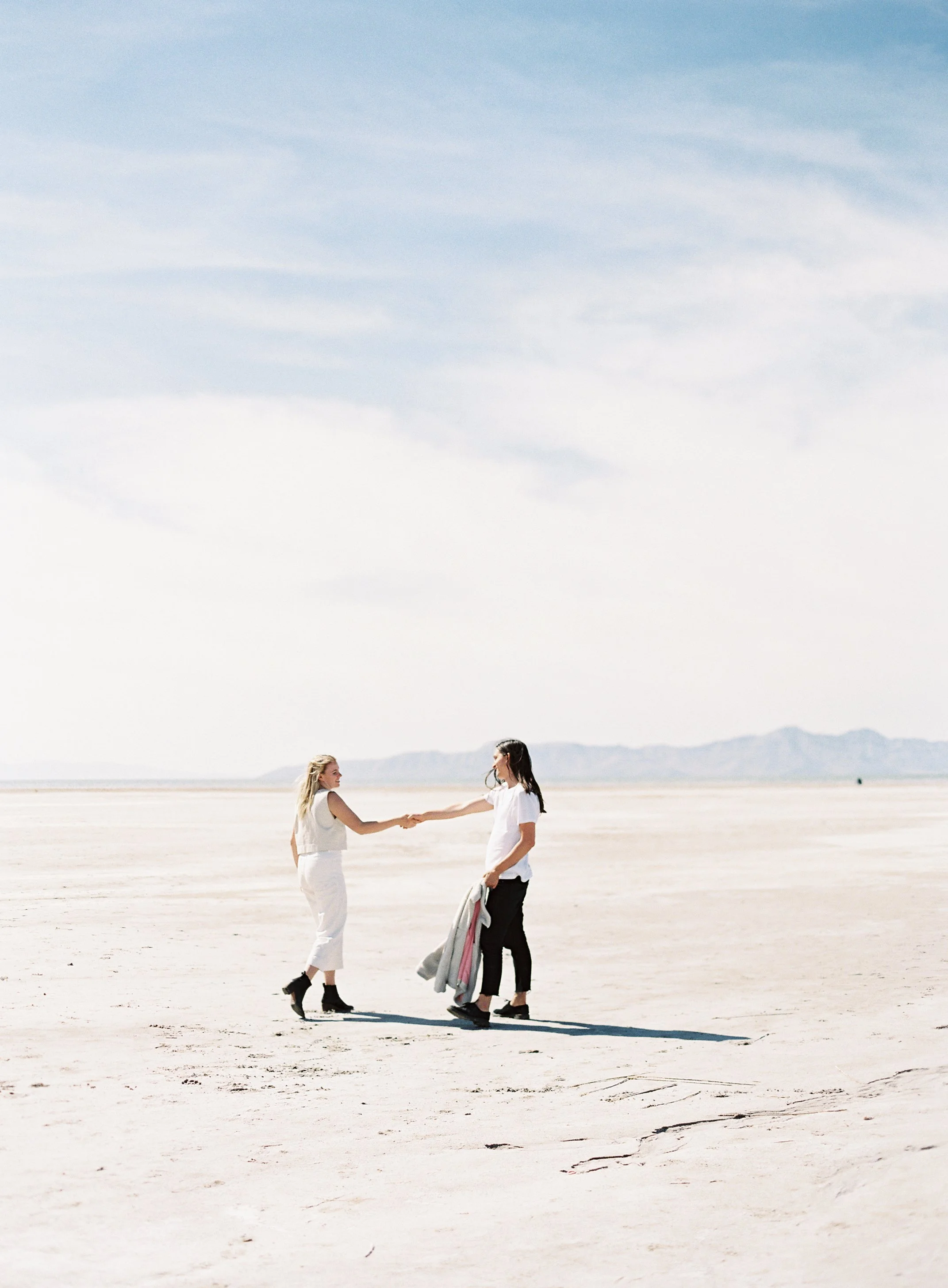 Two women holding hands and smiling at each other on a vast dry, flat landscape with mountains in the background. One woman is dressed in white, the other in black, with some clothing in hand.