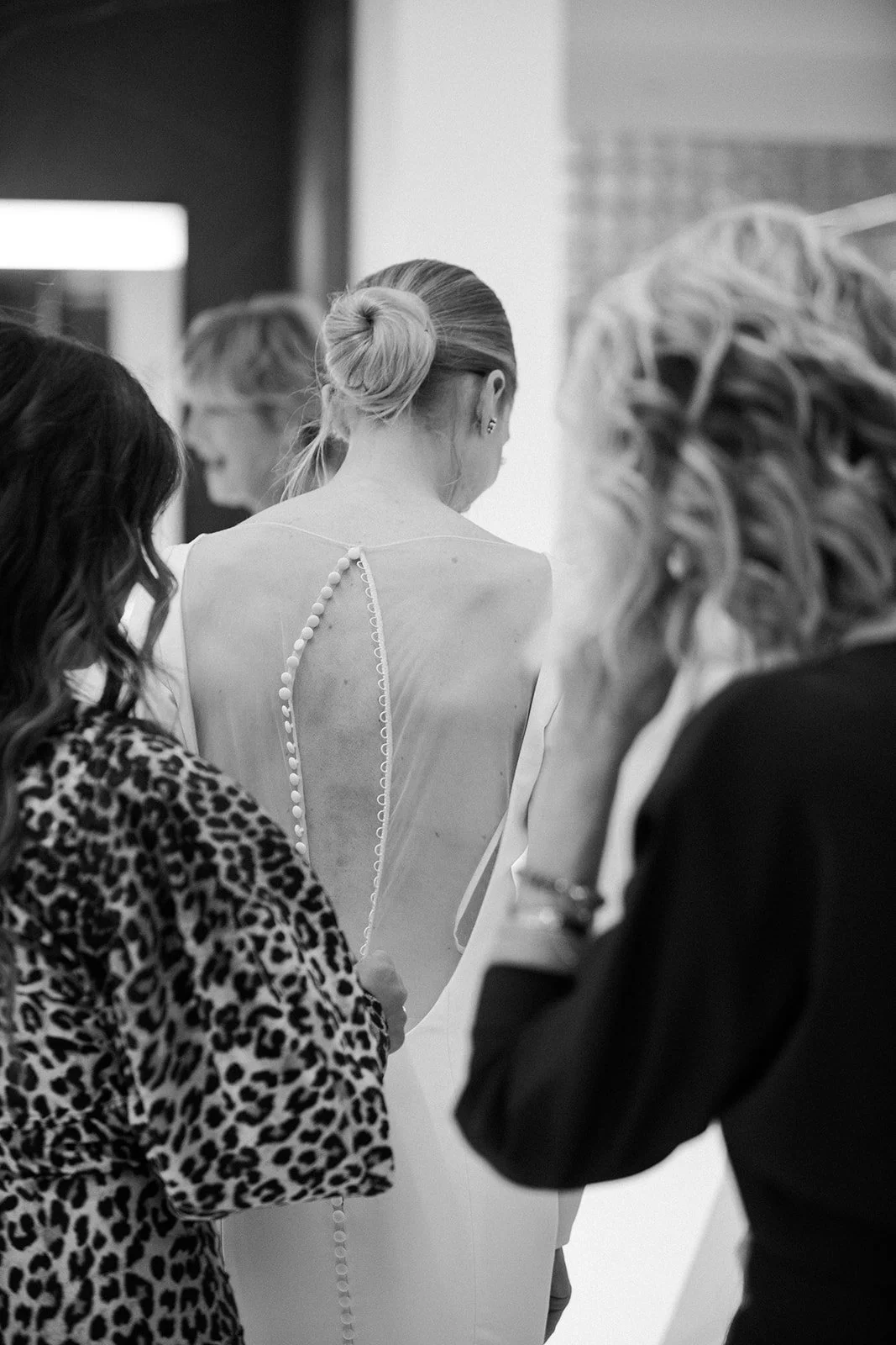 A woman with a bun hairstyle wearing a backless dress with a string of beads, surrounded by three other women, one in a leopard print top, one with curly blonde hair, and another with a ponytail, in a candid backstage setting.