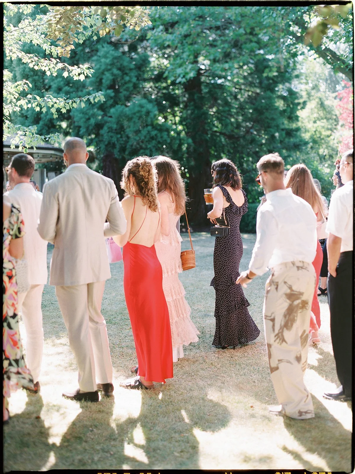 People dressed in formal and semi-formal attire gathering outside on a sunny day, some holding drinks, with lush green trees in the background.